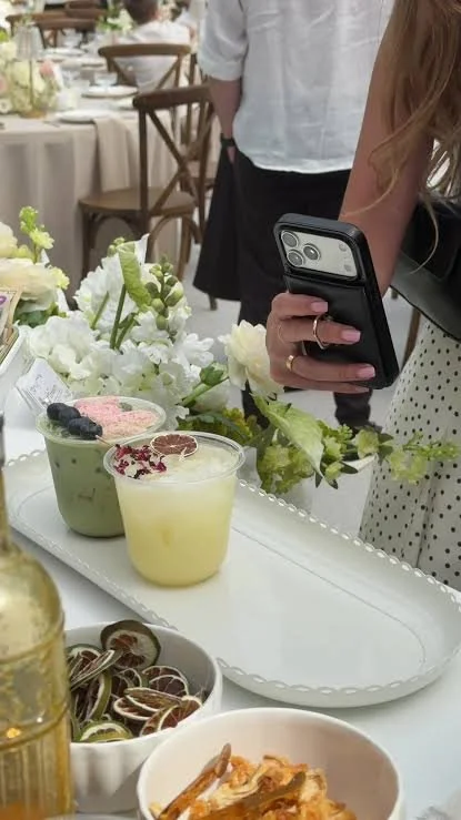 A person holding a smartphone at a table with desserts, including a green dessert with blended fruits, a yellow panna cotta with pomegranate seeds, and a bowl of sliced figs, surrounded by white floral arrangements and other food items.
