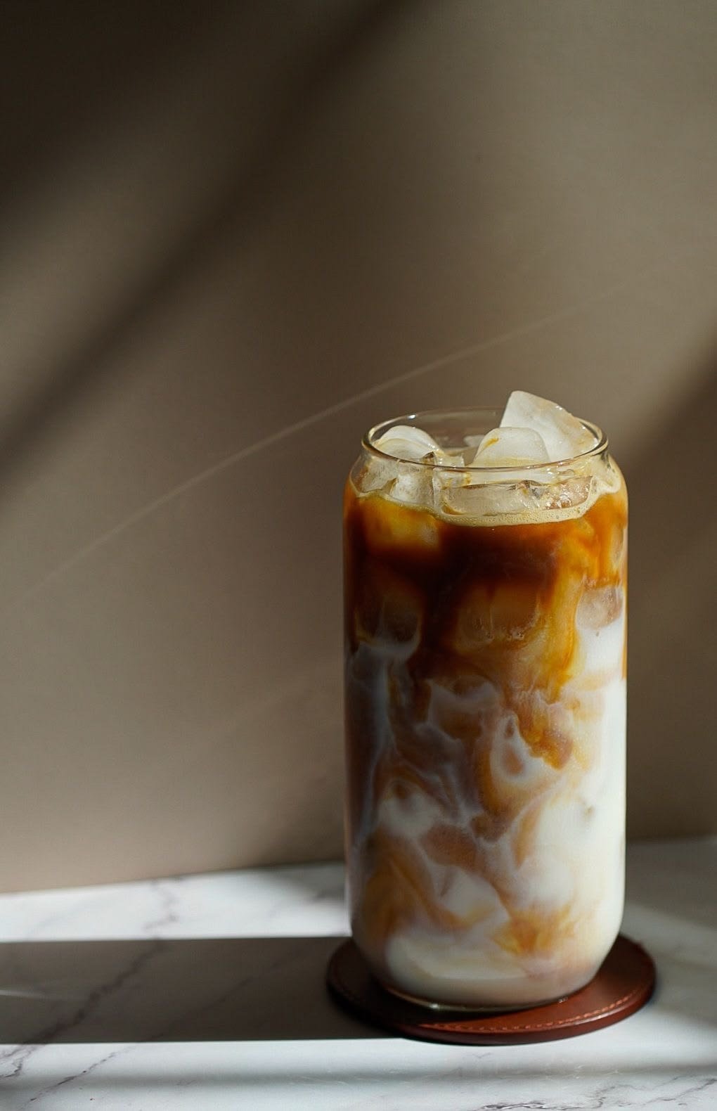 A tall glass of iced coffee with milk and caramel swirls, filled with ice cubes, sitting on a white marble surface with a neutral background.