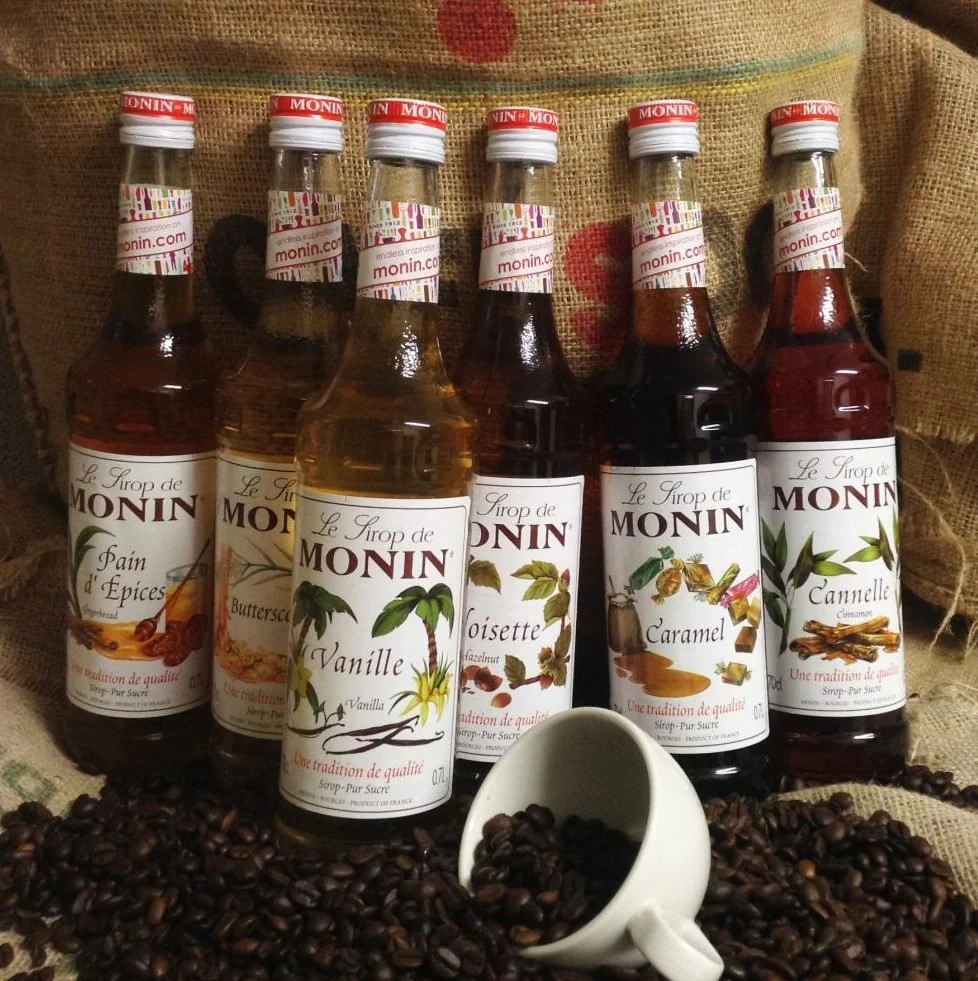 Six bottles of Monin flavored syrup, including vanilla, caramel, cinnamon, butterscotch, peacock, and hazelnut, surrounded by coffee beans and a white espresso cup filled with coffee beans. The bottles are arranged on a fabric surface with a burlap backdrop.