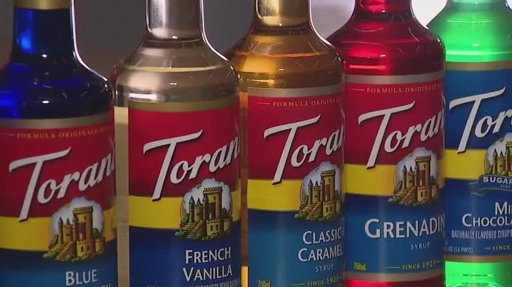 Five bottles of Torani flavored syrup lined up, each with a different flavor label: Blue, French Vanilla, Classic Caramel, Grenadine, and a green-labeled flavor.