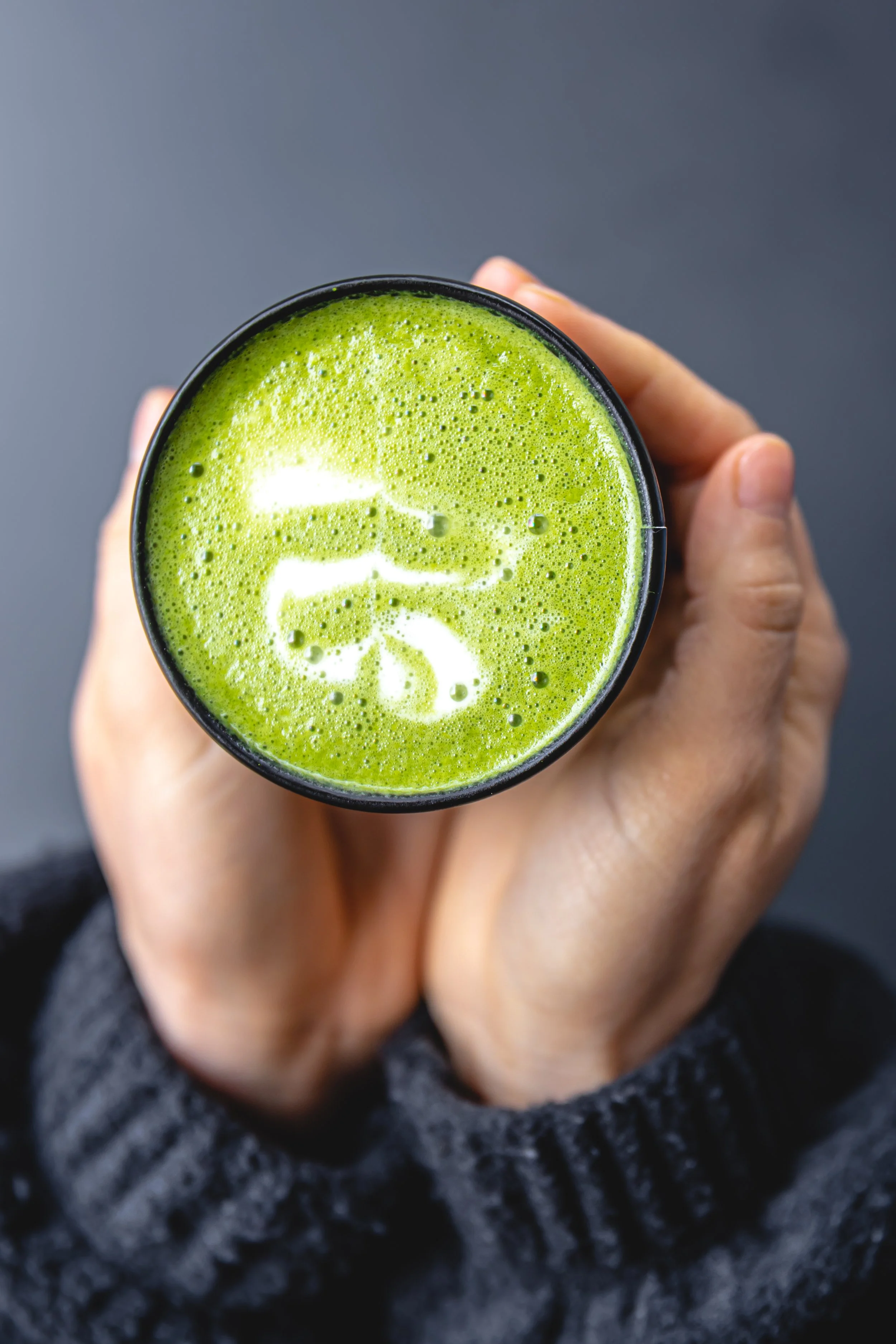 Top view of a person holding a black mug filled with green matcha latte topped with latte art in the shape of a leaf.