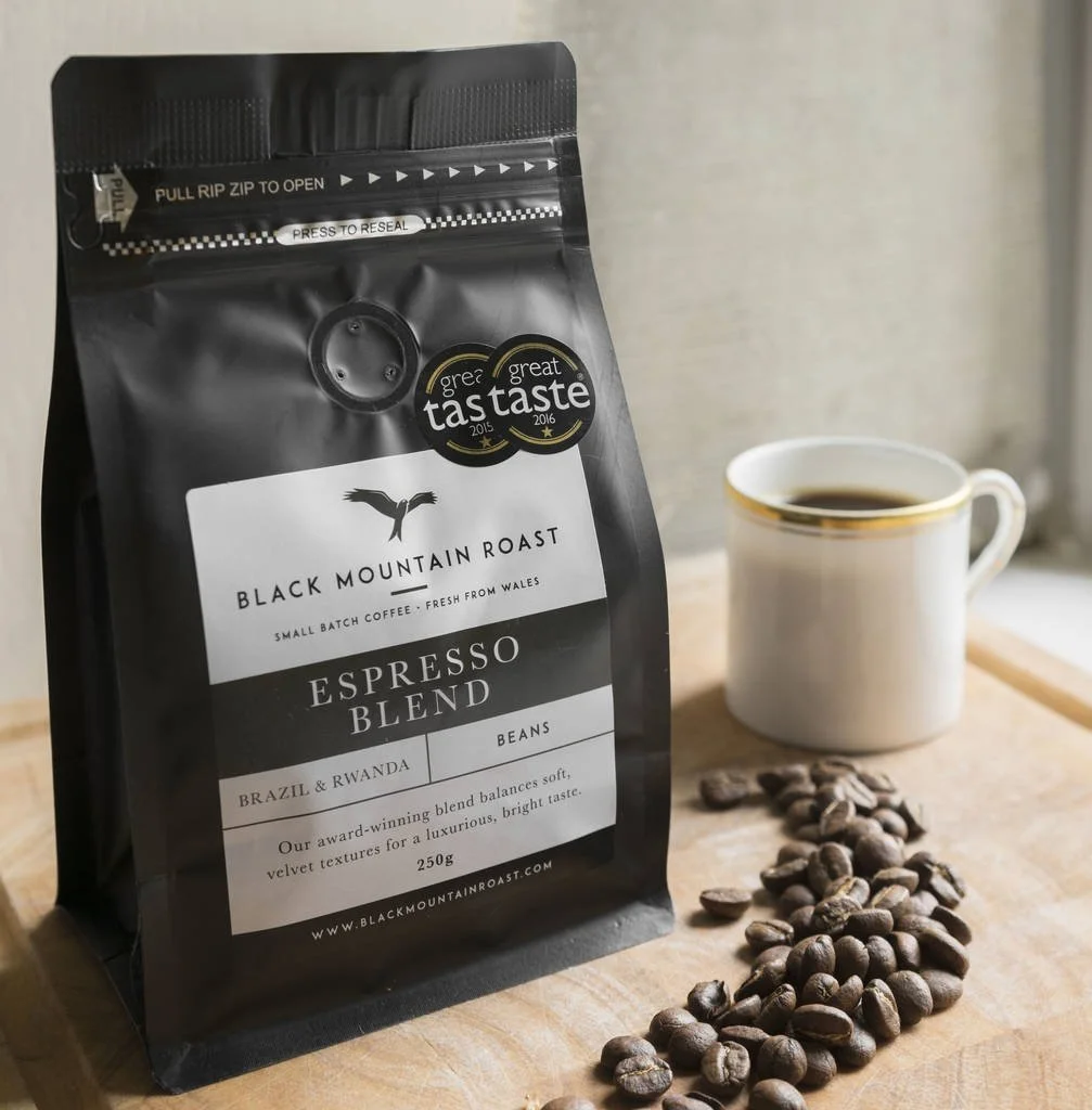 A bag of Black Mountain Roast Espresso Blend coffee beans next to a white mug filled with coffee and scattered coffee beans on a wooden surface.