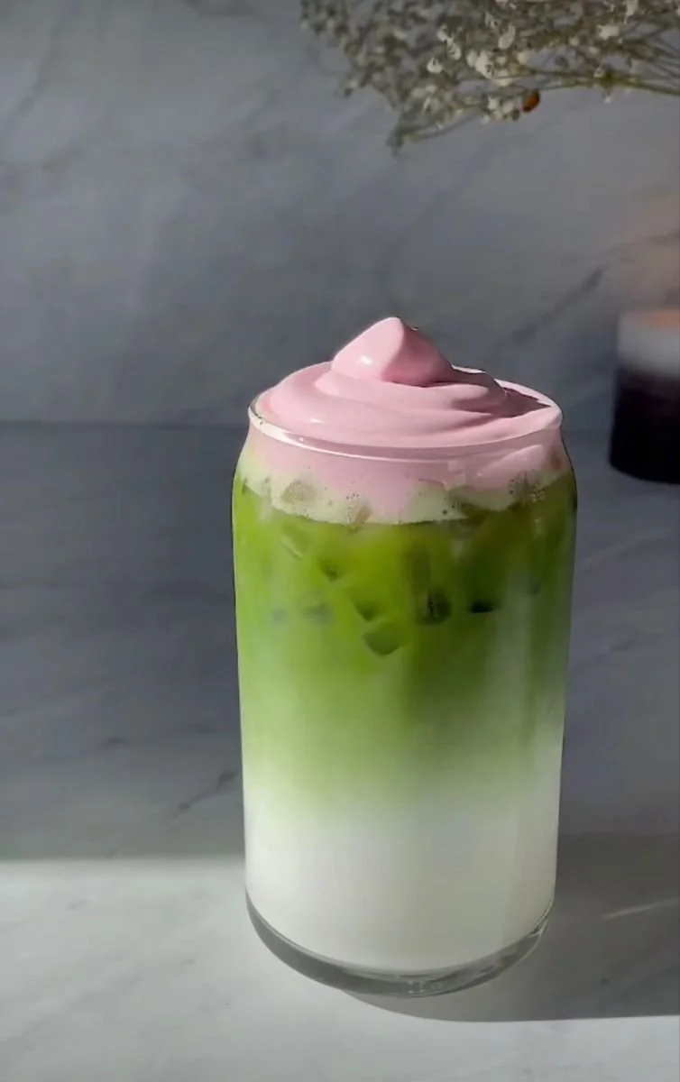 Layered beverage in a clear glass with green tea, milk, and pink whipped topping.