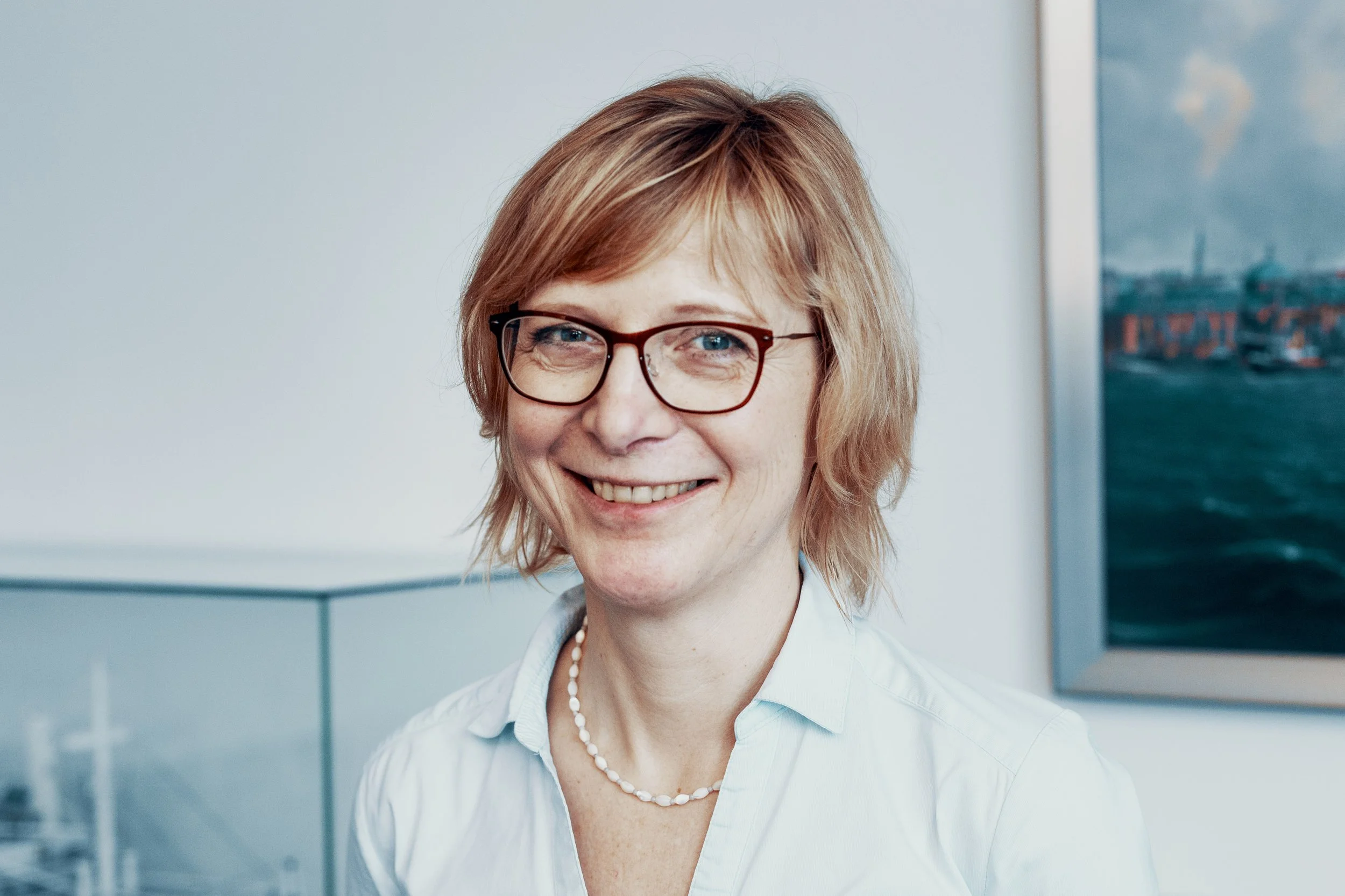 Ein Porträt einer Frau mit kurzen roten Haaren, Brille und einem weißen Hemd, die vor einem modernen Hintergrund steht.