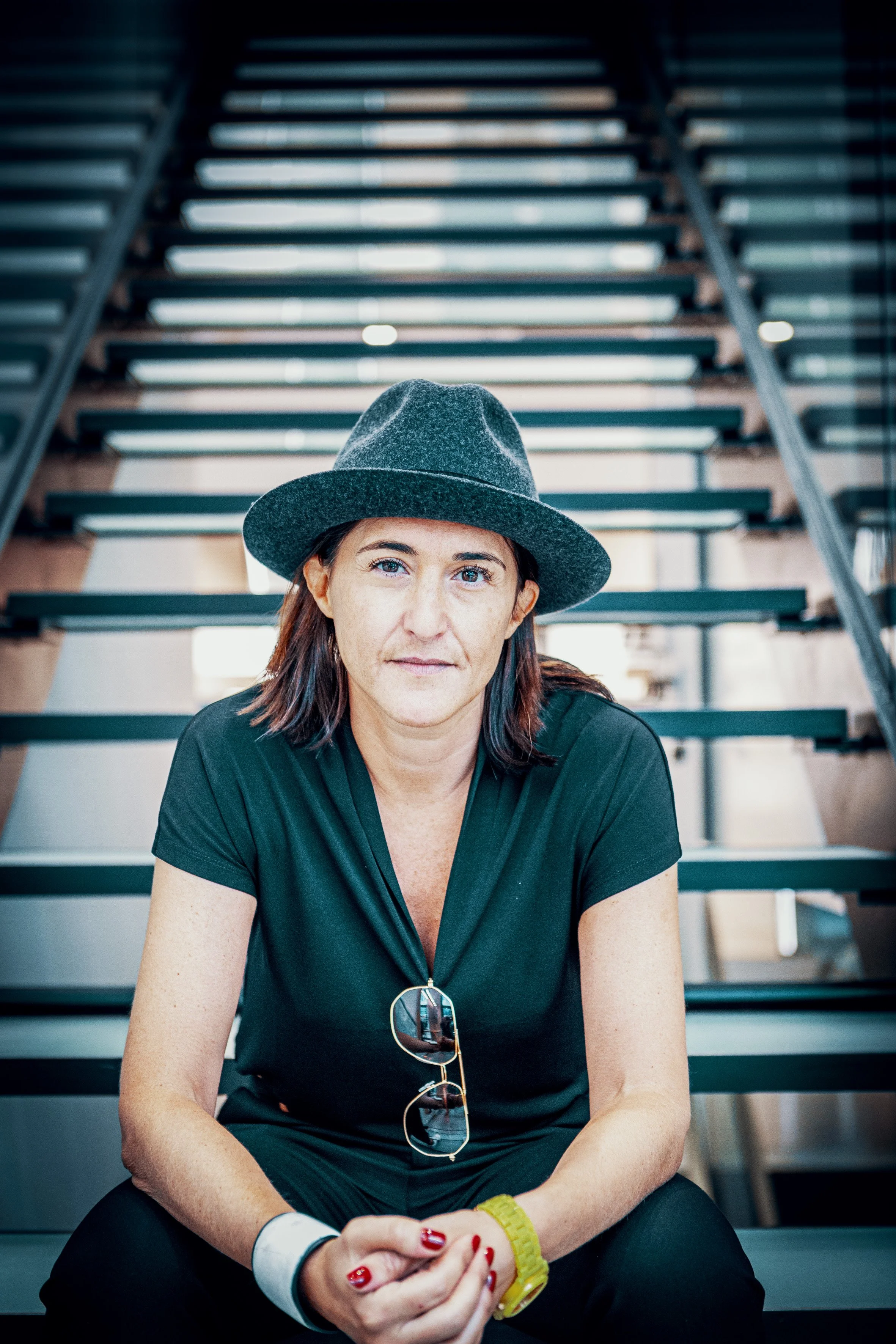 Junge Frau mit grauem Hut und schwarzen Kleid sitzt auf einer Treppe, schaut in die Kamera, trägt eine Sonnenbrille und hat rote Fingernägel.