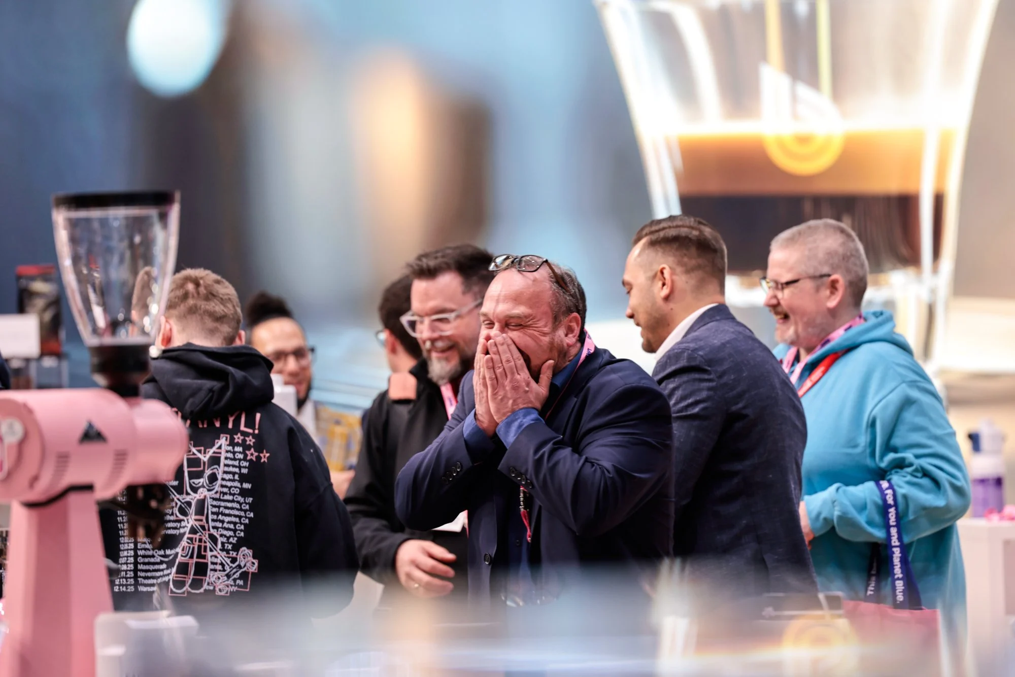 Menschen lachen und haben Spaß bei einem gemeinsamen Ereignis, sitzen an einem Tisch mit Getränken, im Hintergrund eine Beleuchtung und eine große Tafel
