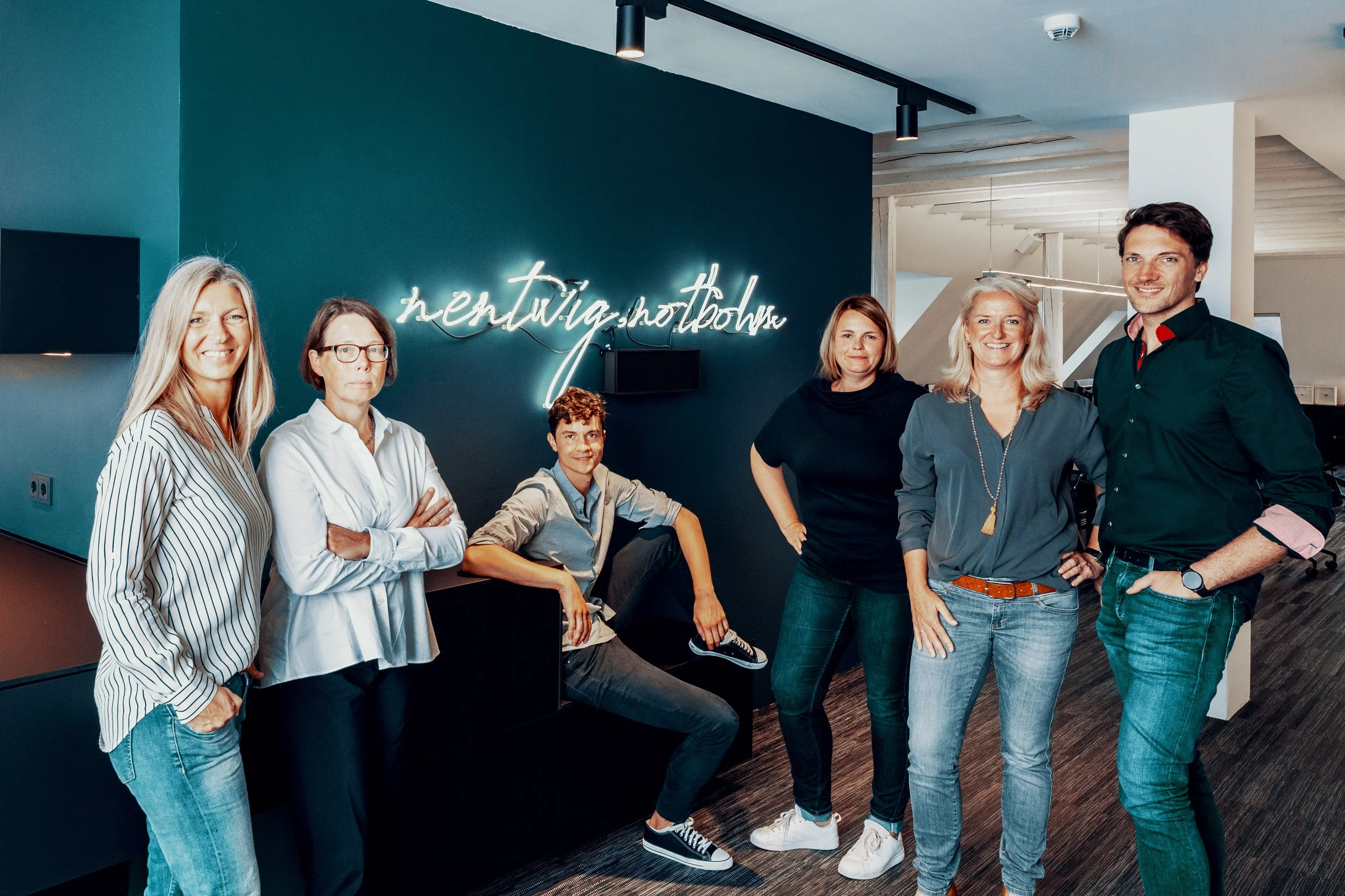 Eine Gruppe von sieben Menschen posiert in einem modernen Büro vor einer dunklen Wand mit einem Neon-Zeichen, das 'hentwig shooting' liest.