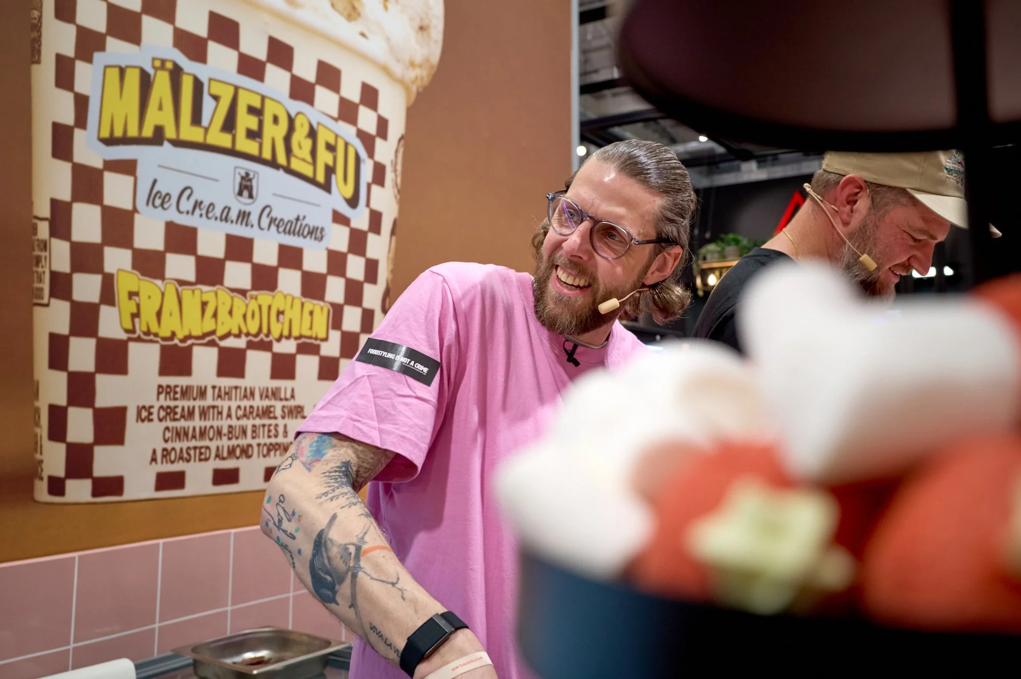 Zwei Männer lächeln und sprechen, während sie in einem Laden stehen, mit einem großen Eiscreme Produkt in der Nähe im Vordergrund. Der Hintergrund zeigt eine Wand mit einem Werbebanner für Eiscreme, auf dem "MALZER & FU" und eine Eistüte mit verschie