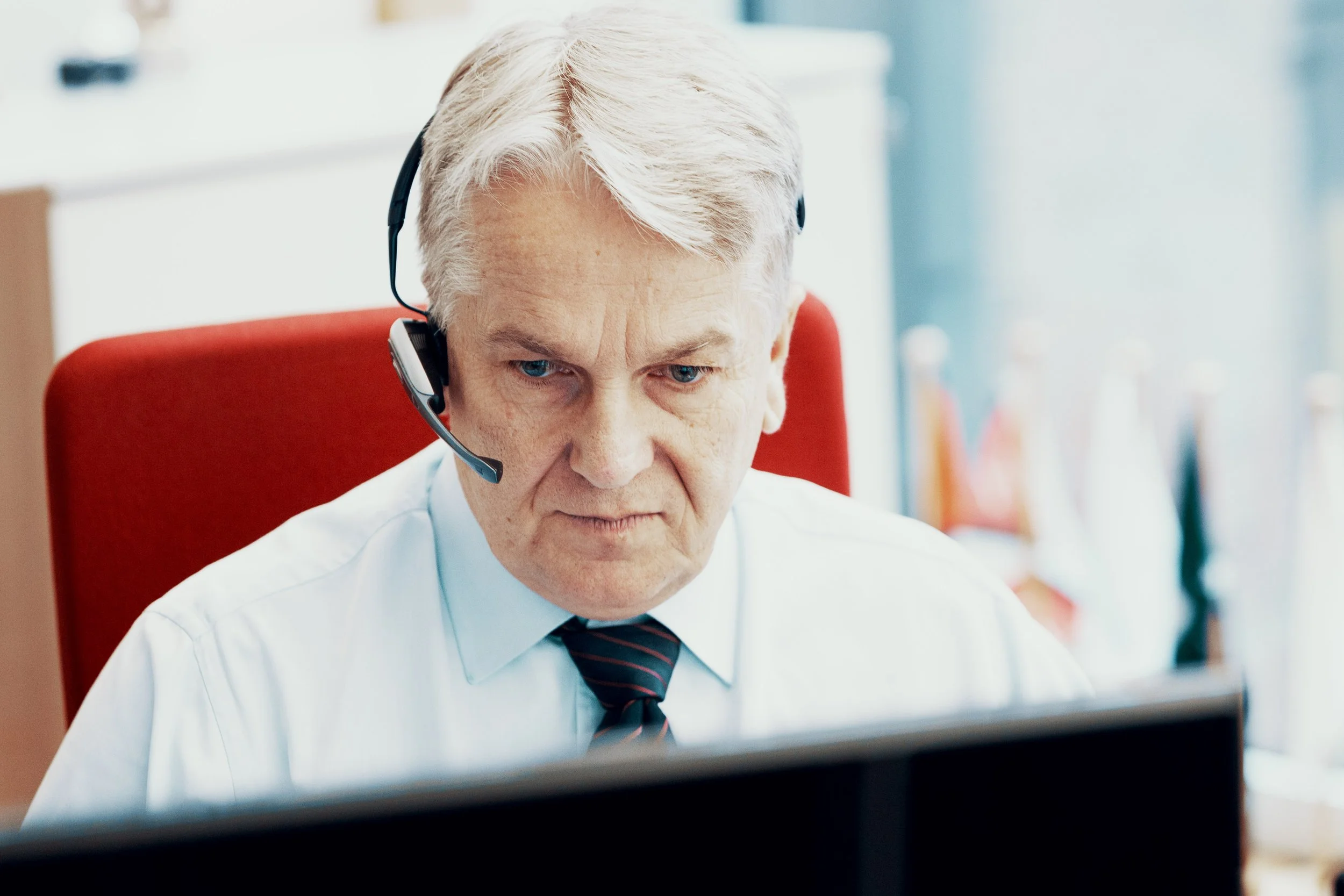 Ein älterer Mann mit weißen Haaren trägt ein Headset und arbeitet an einem Computer im Büro.