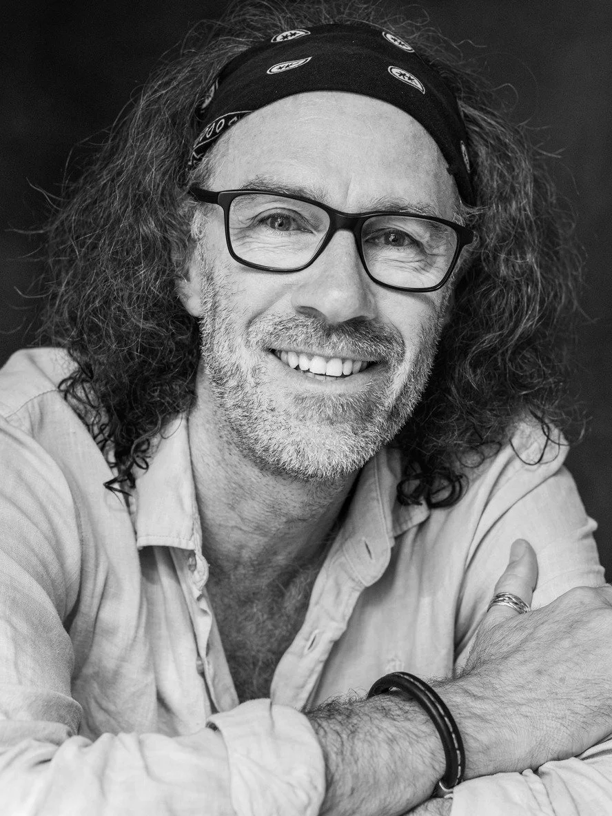 Mann mit langen, lockigen Haaren, Brille und einem Bandana, lächelnd, in einem Hemd, schwarze und weiße Fotografie
