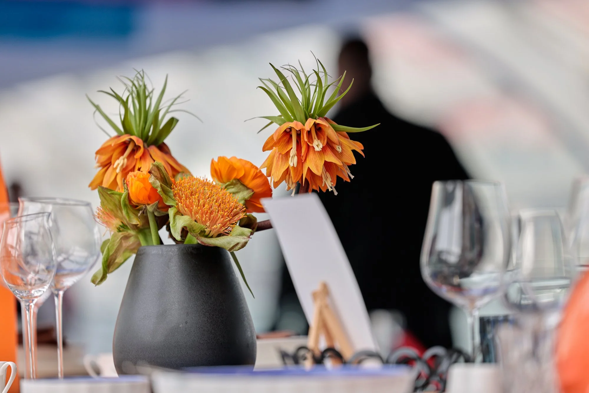Blumen in einer grauen Vase auf einem Tisch, im Hintergrund unscharfe Gläser und ein Mensch.