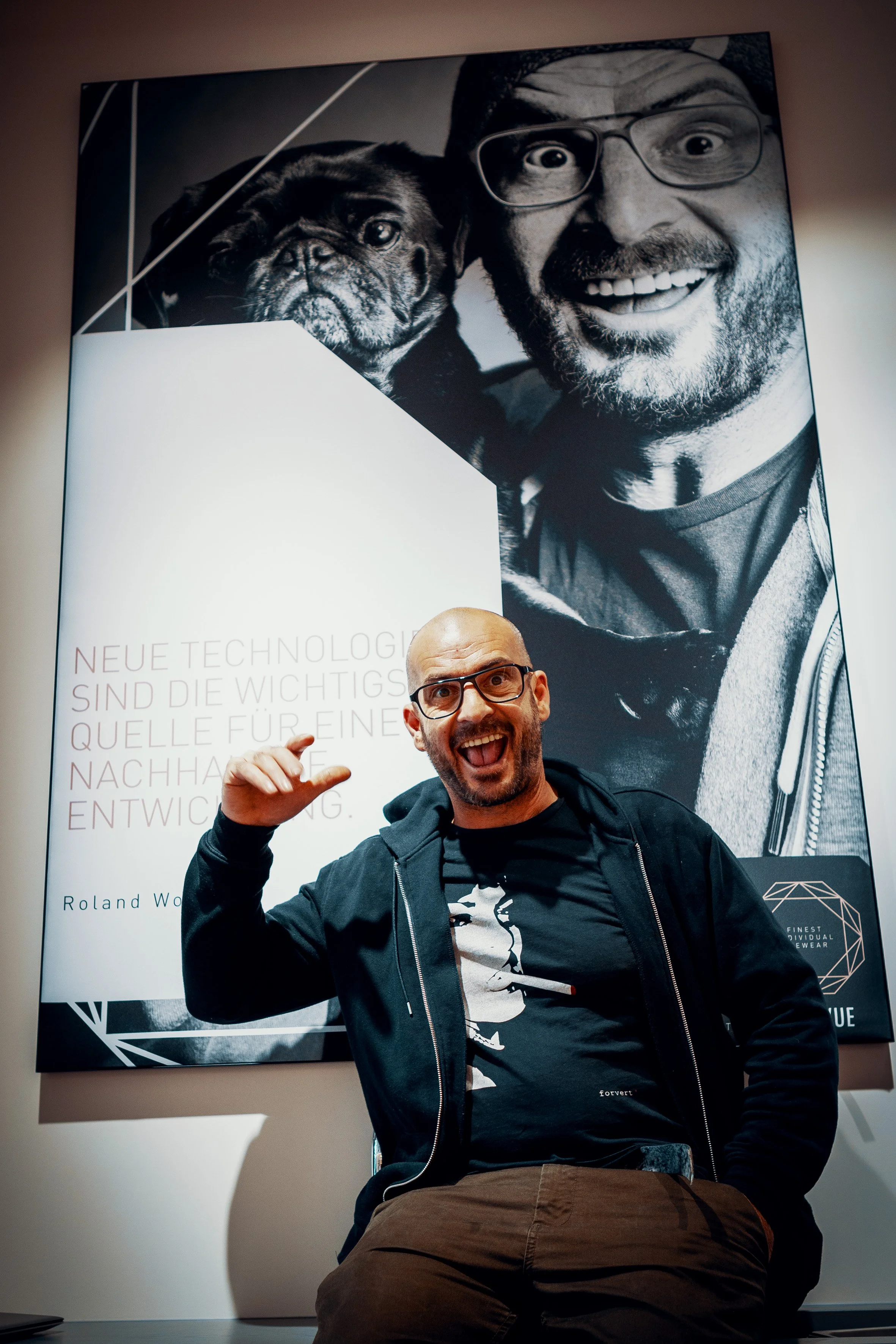 Junger Mann mit Brille, lachend und mit Hand am Kopf, sitzt vor einem großen Schwarz-Weiß-Poster eines Mannes mit Brille und Hund, im Hintergrund.