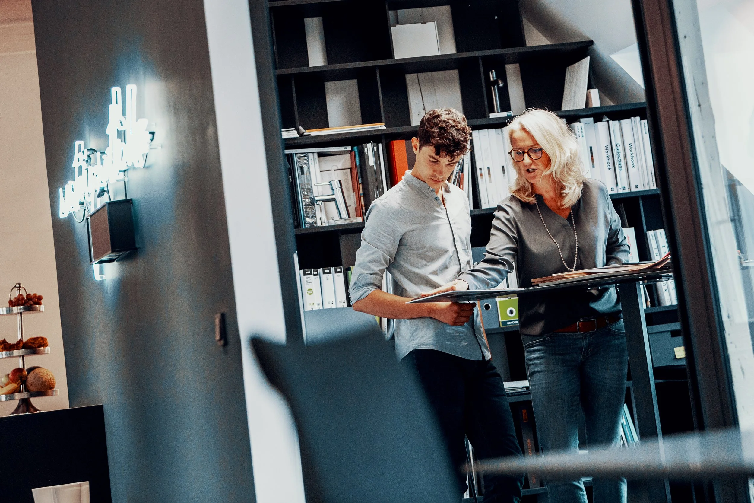 Zwei Frauen stehen in einem modernen Büro, schauen auf Dokumente und diskutieren. Im Hintergrund Ratgeberregale und vor einem Fenster.