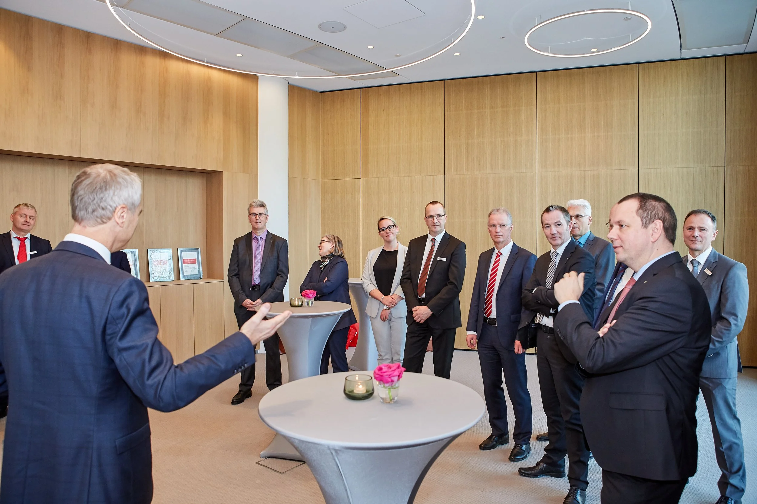 Eine Gruppe von Geschäftsleuten in Anzügen bei einem Meeting in einem modernen, holzverkleideten Raum, wobei die meisten den Sprecher am runden Stehtisch beobachten.