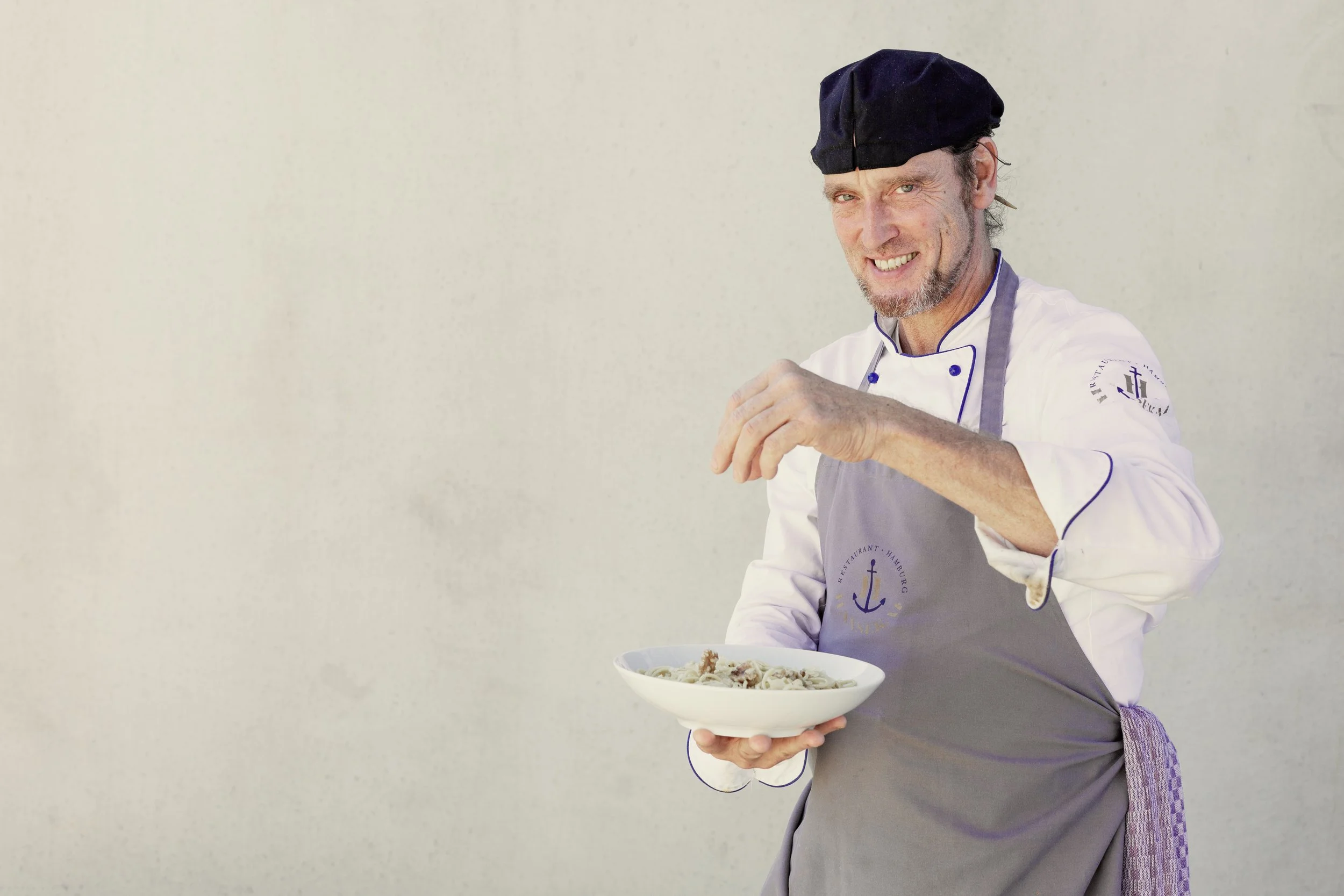Ein freundlicher männlicher Koch in uniform und Haube hält eine Schale mit Pasta und schaut in die Kamera.