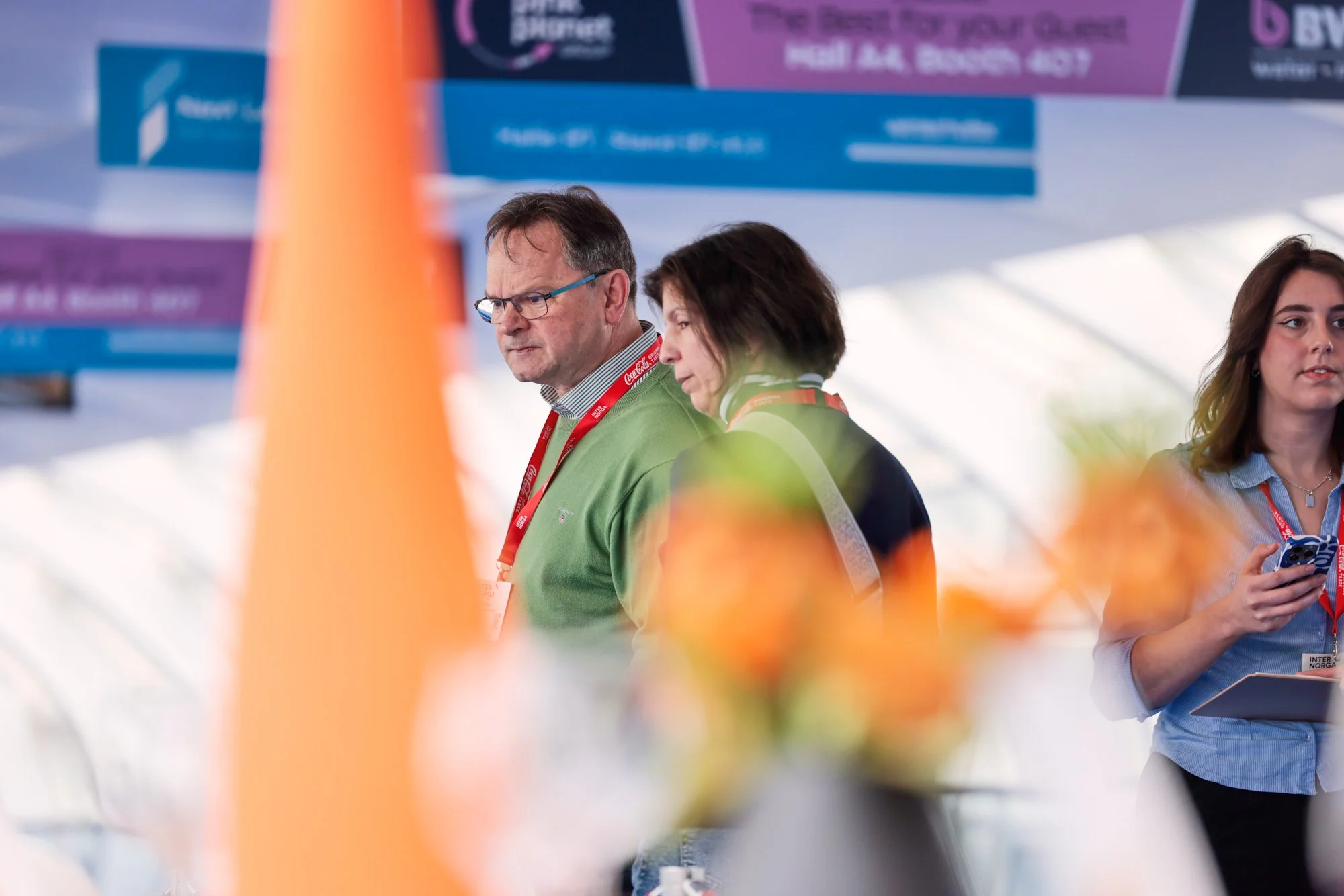 Menschen an einer Messe oder Konferenz, teilweise unscharf im Vordergrund, im Hintergrund Banner mit Text und einem blauen Himmel sichtbar.