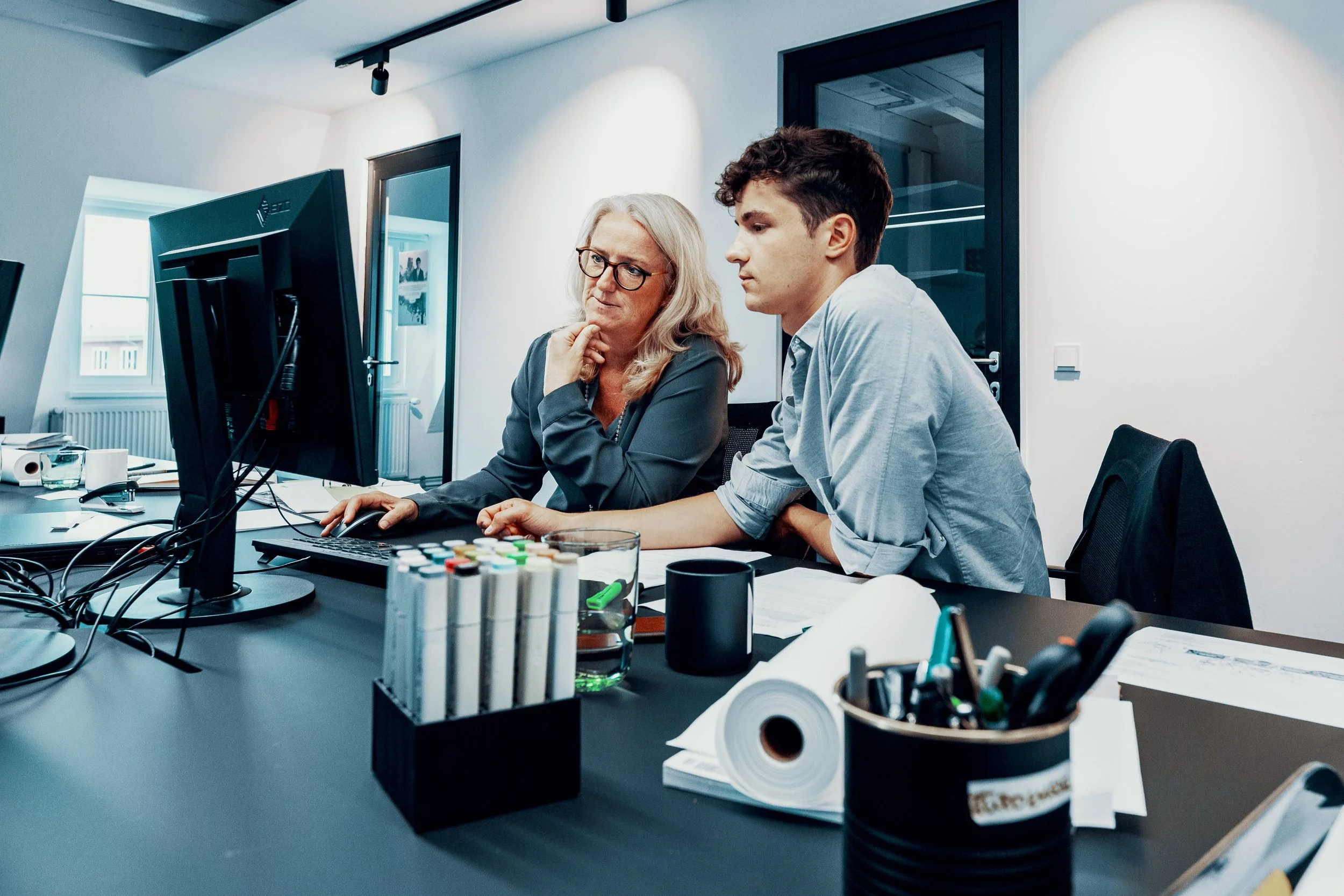 Zwei Personen, eine ältere Frau mit Brille und eine jüngere Person, sitzen in einem modernen Büro vor einem Computer und diskutieren. Das Büro ist hell erleuchtet, mit weißen Wänden und Fenstern im Hintergrund.