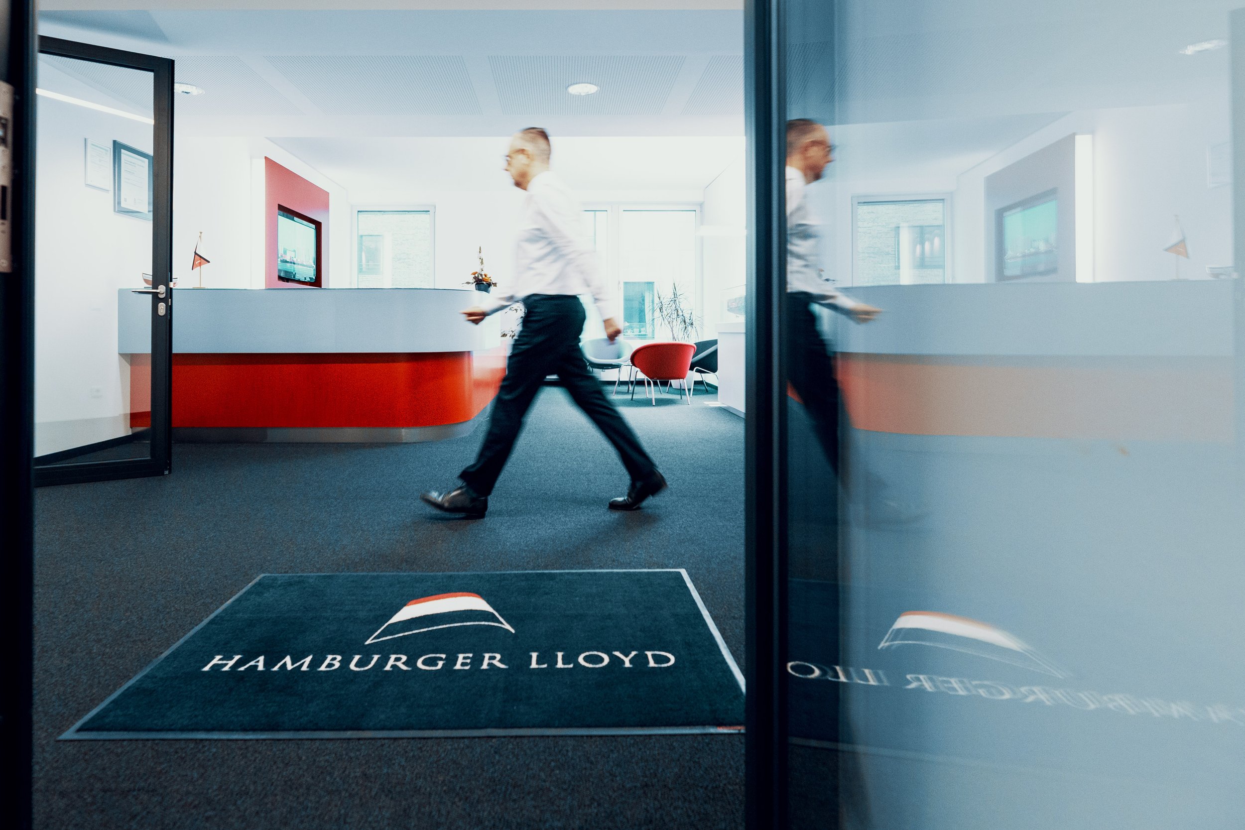 Ein Mann in Businesskleidung läuft über den Eingangsbereich des Büros der Hamburger Lloyd, sichtbar durch die Glastür. Im Hintergrund befindet sich eine moderne Empfangsbereich mit red-weißer Theke, Sitzecke und Fernseher.