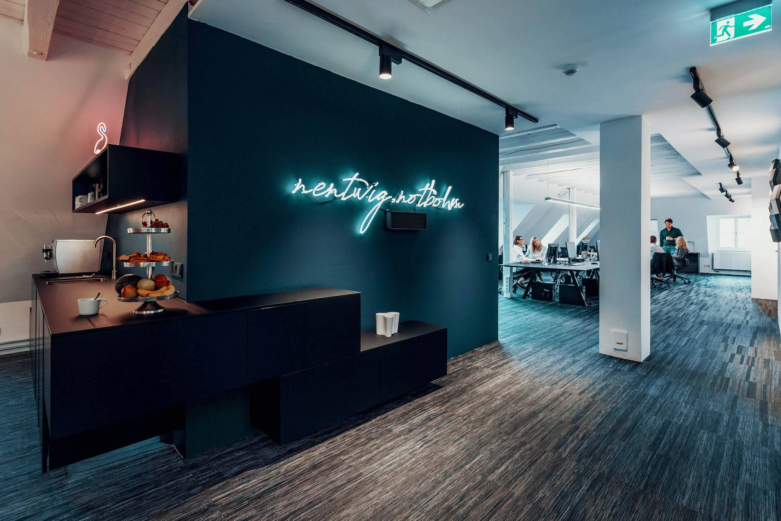Moderne Bürosoft-Lounge mit Kaffee und kleinen Snacks, blauer Wand mit Neon-Schriftzug 'hentwig.notbohne' und Mitarbeitern am Arbeitsplatz im Hintergrund.