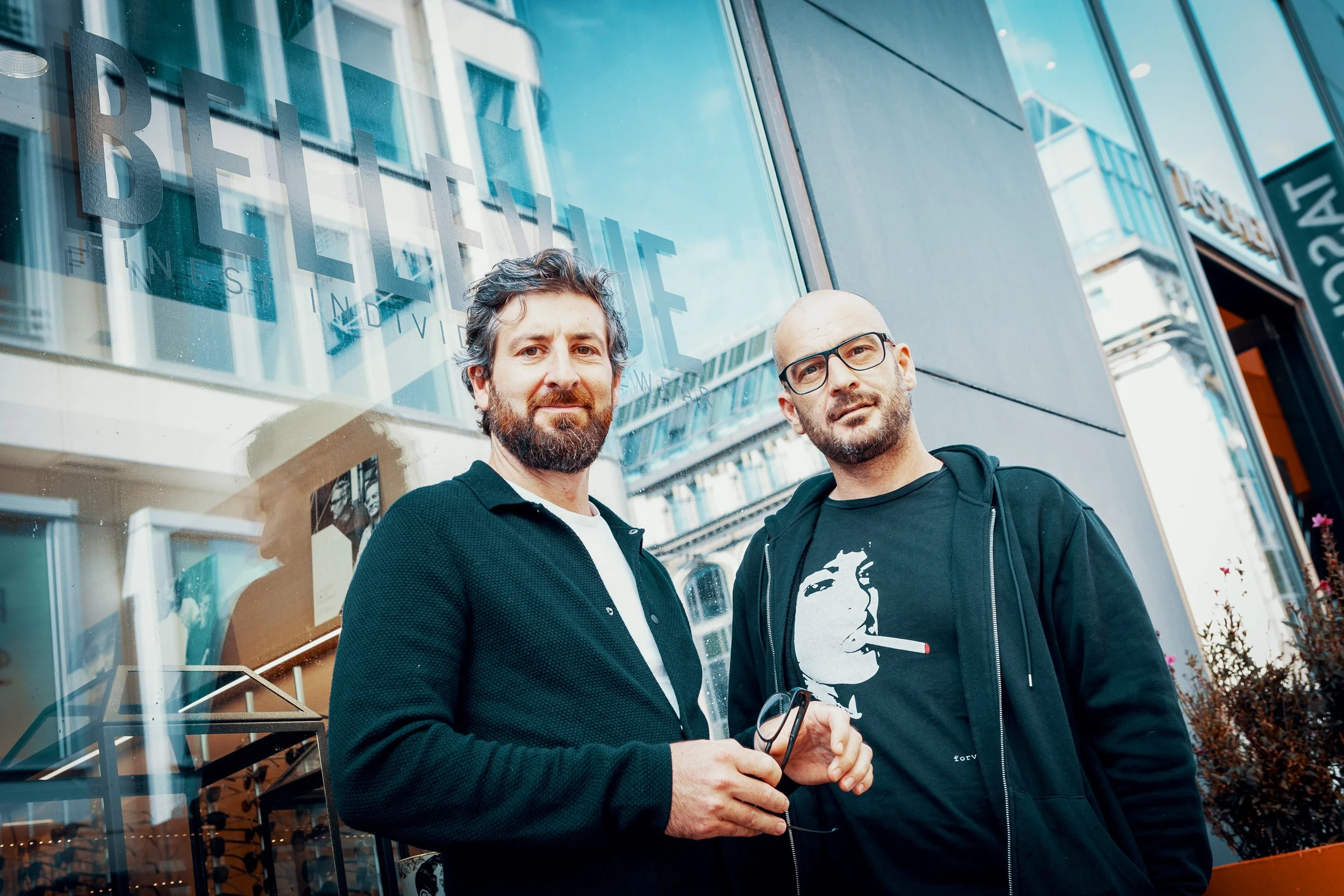 Zwei Männer stehen vor einem großen Fenster in einem modernen Gebäude, das reflektiert. Einer trägt eine schwarze Jacke und hat einen Bart, der andere trägt eine Brille und ein schwarzes T-Shirt mit einem weißen Portrait. Sie scheinen sich die Hand z