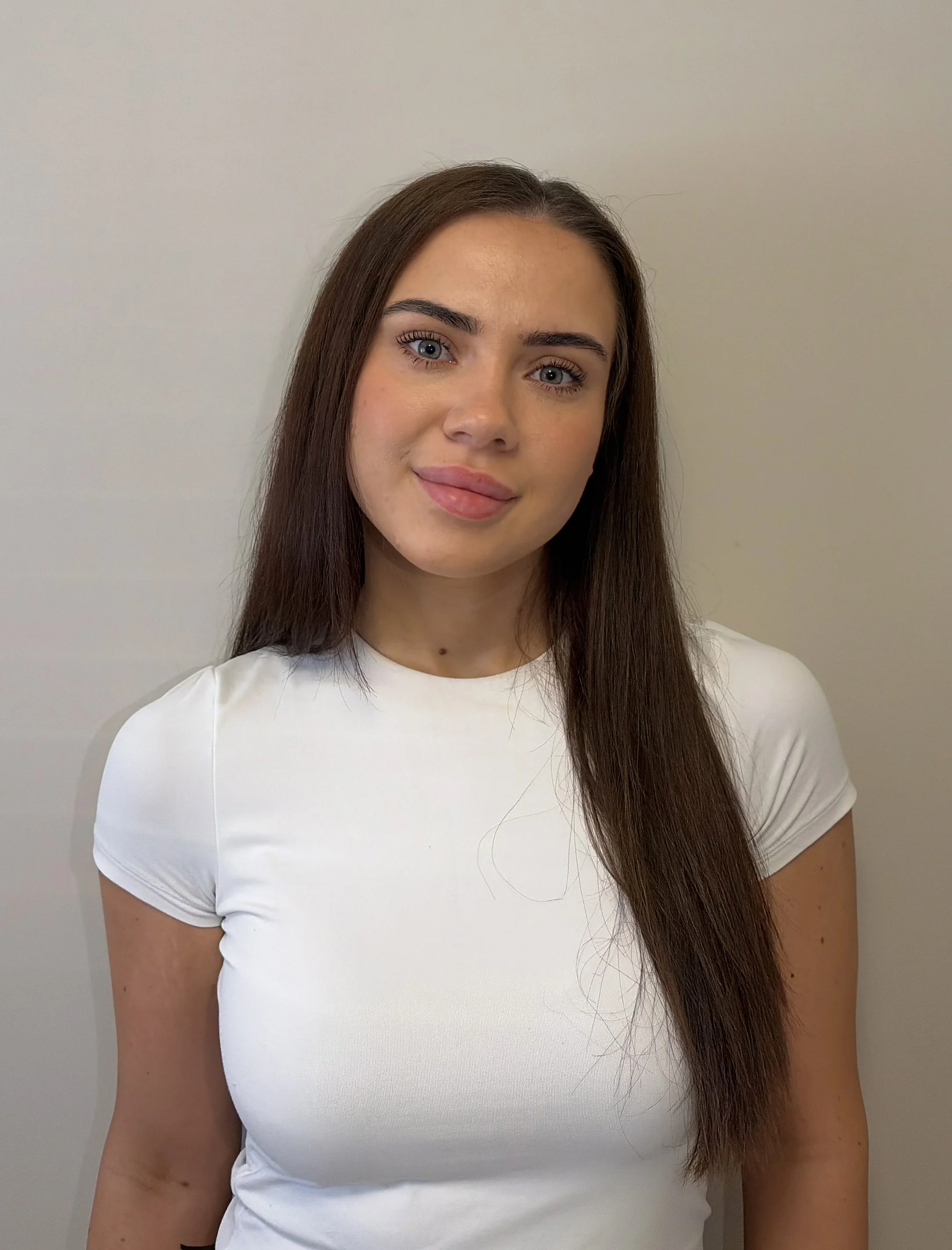 A young woman with long brown hair and blue eyes, wearing a white shirt, standing against a plain gray wall.
