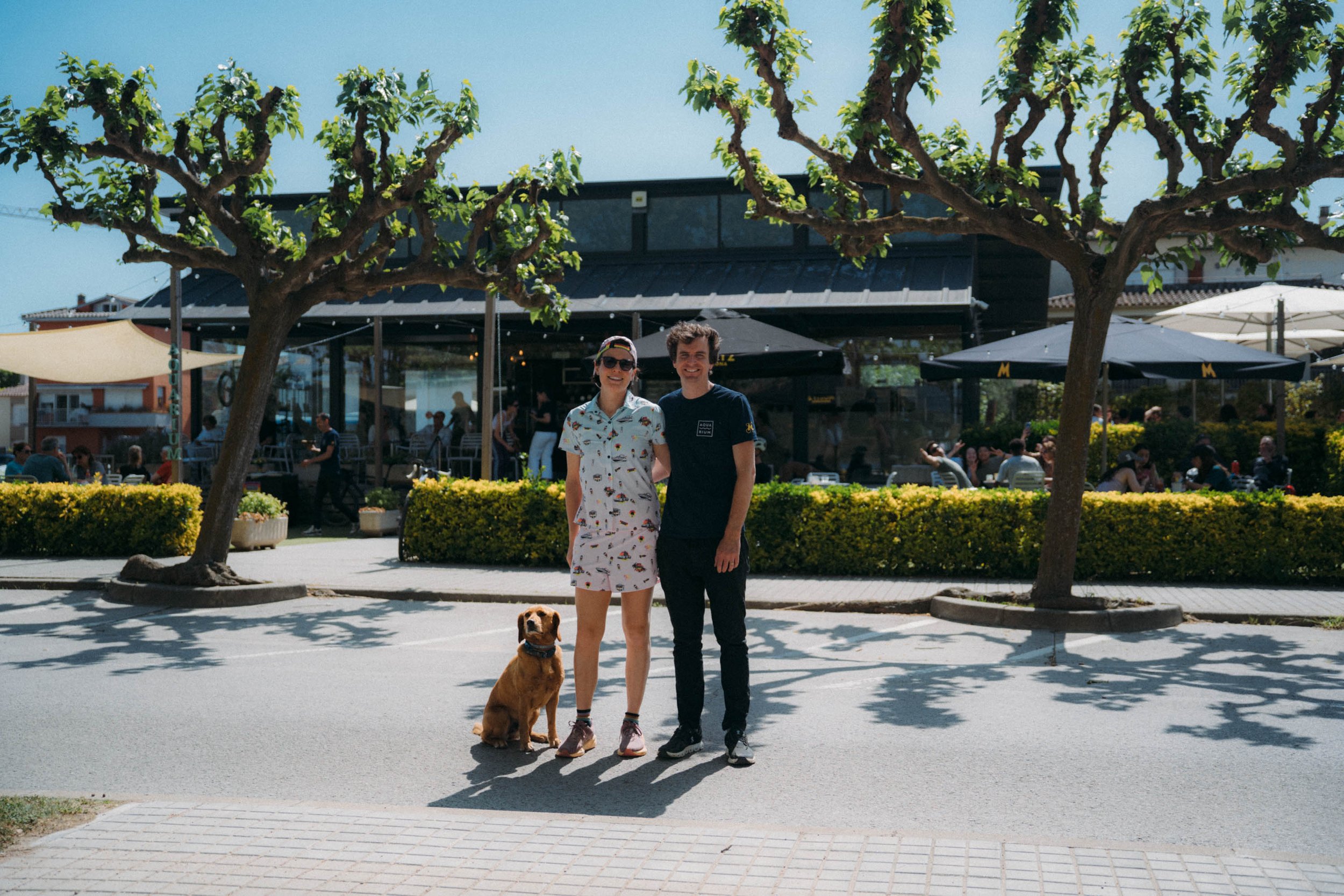 Two people smiling and standing with a dog on a sunny day in an outdoor area with trees, bushes, and a building with outdoor seating.