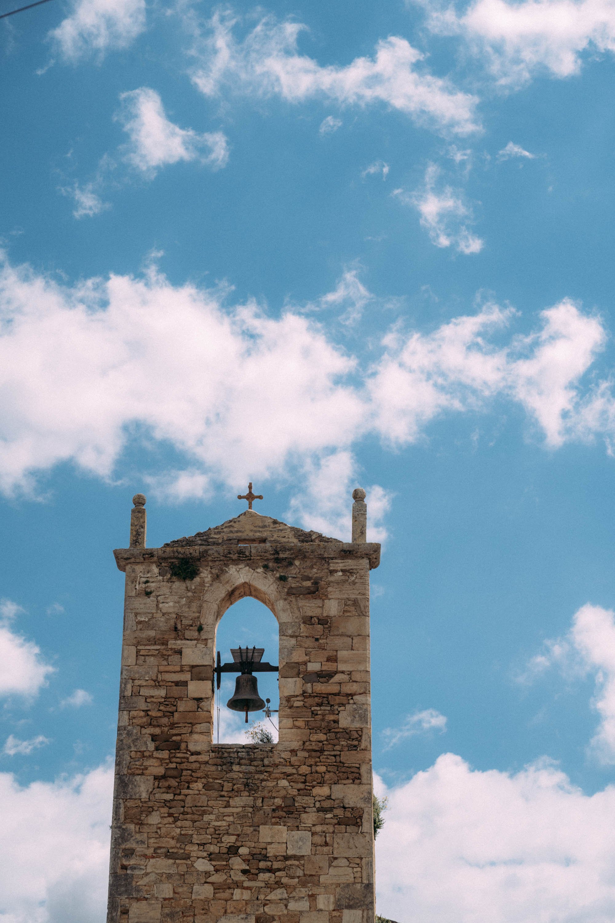Stone church bell tower with a cross on top, set against a blue sky with scattered white clouds.