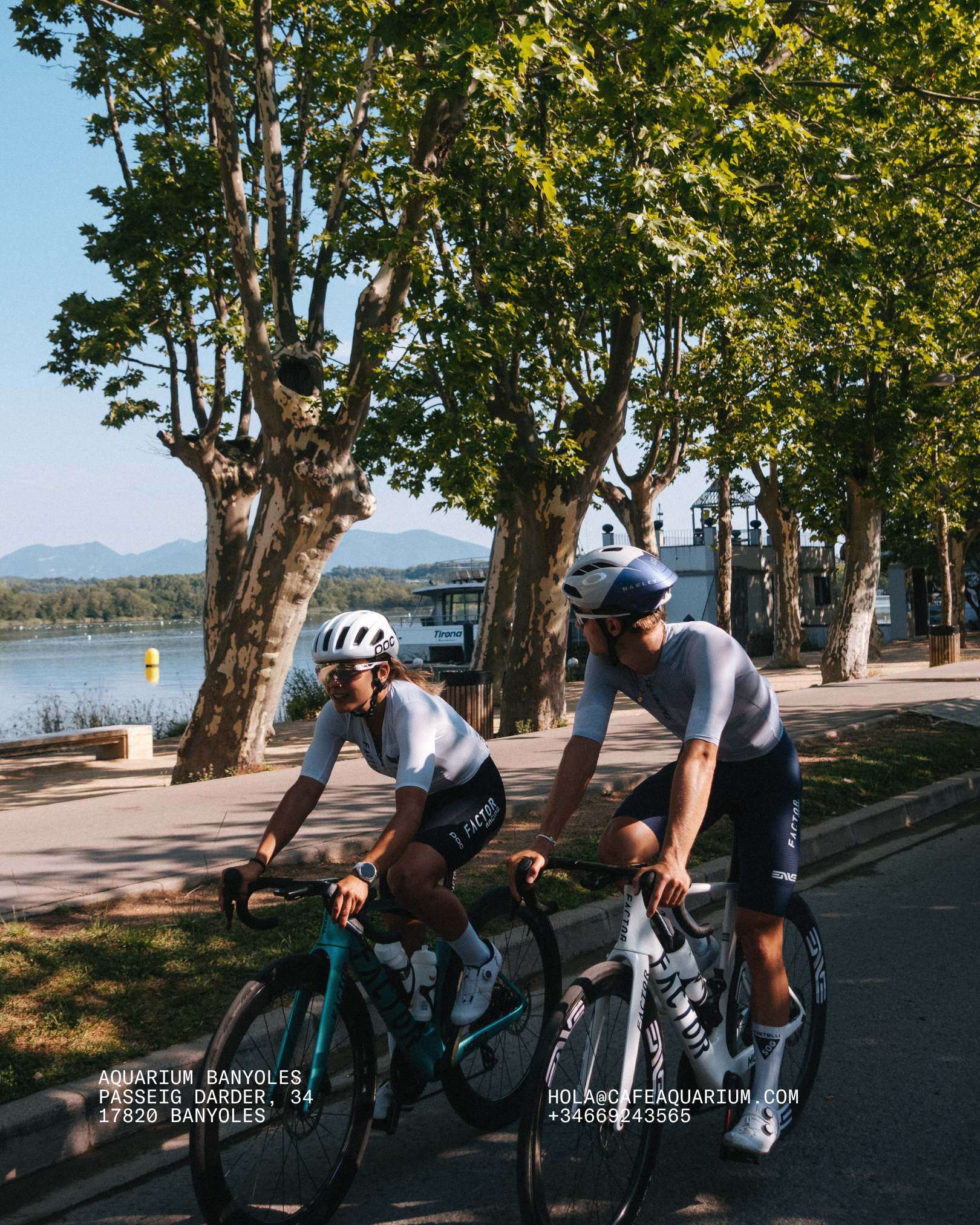 Cyclists riding bikes along a waterfront with trees and a boat in the background on a sunny day.
