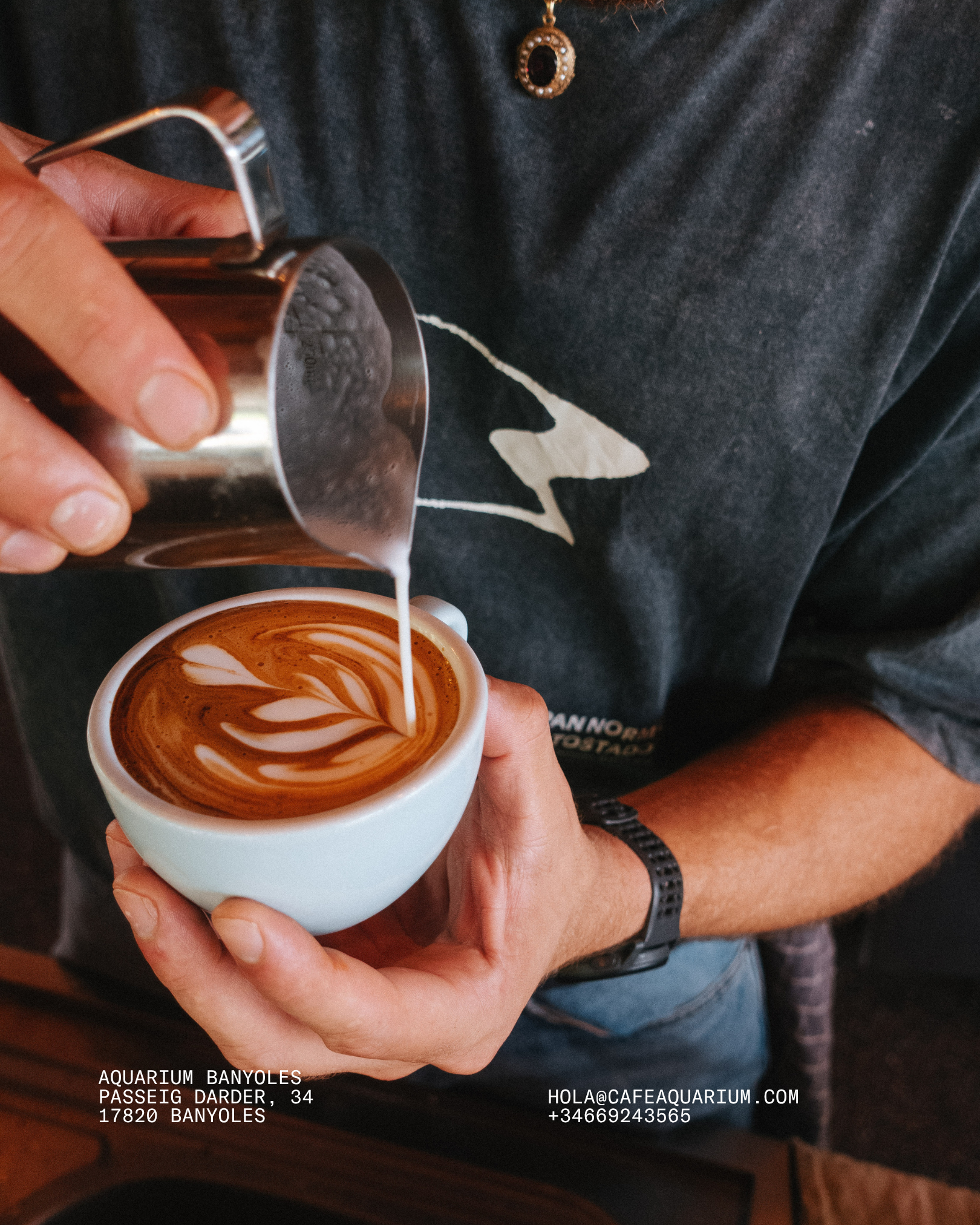 A person is pouring milk into a cup of coffee with latte art. The person is wearing a dark gray shirt and a black wristwatch. There is contact information and location printed on the image, indicating it is from an aquarium cafe in Banyoles, Spain.