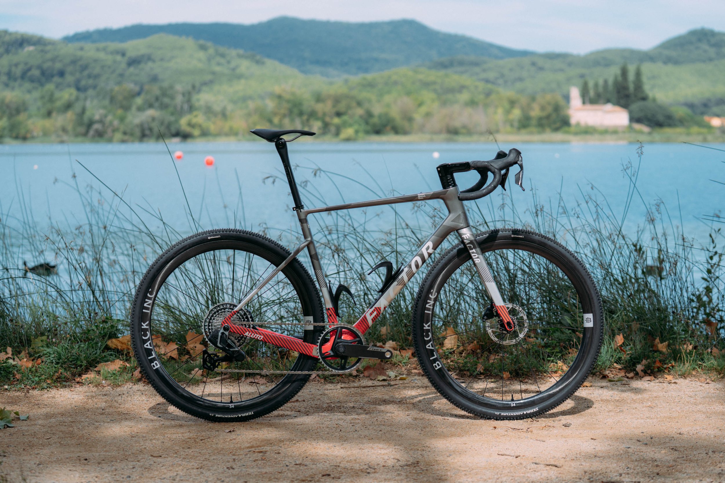 A mountain bike with a red and black frame parked on a dirt path near a calm lake, with lush green hills and a building visible in the background.