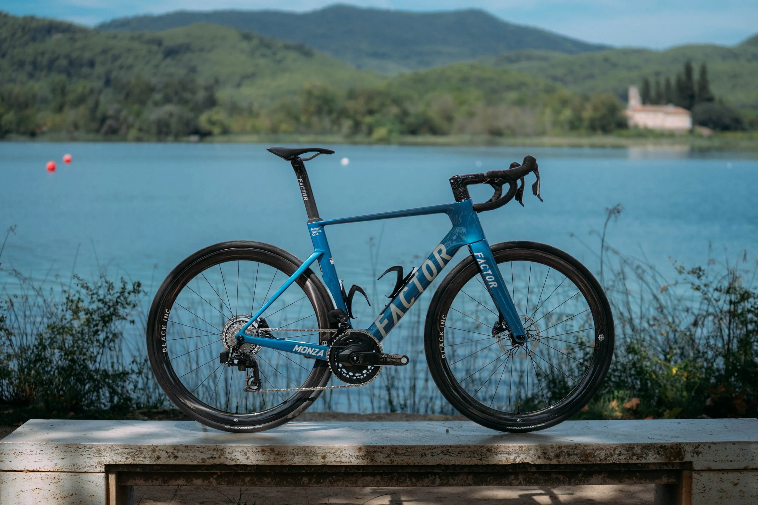 A blue bicycle with black tires and a black seat, resting on a white platform near a body of water with a mountainous landscape in the background.