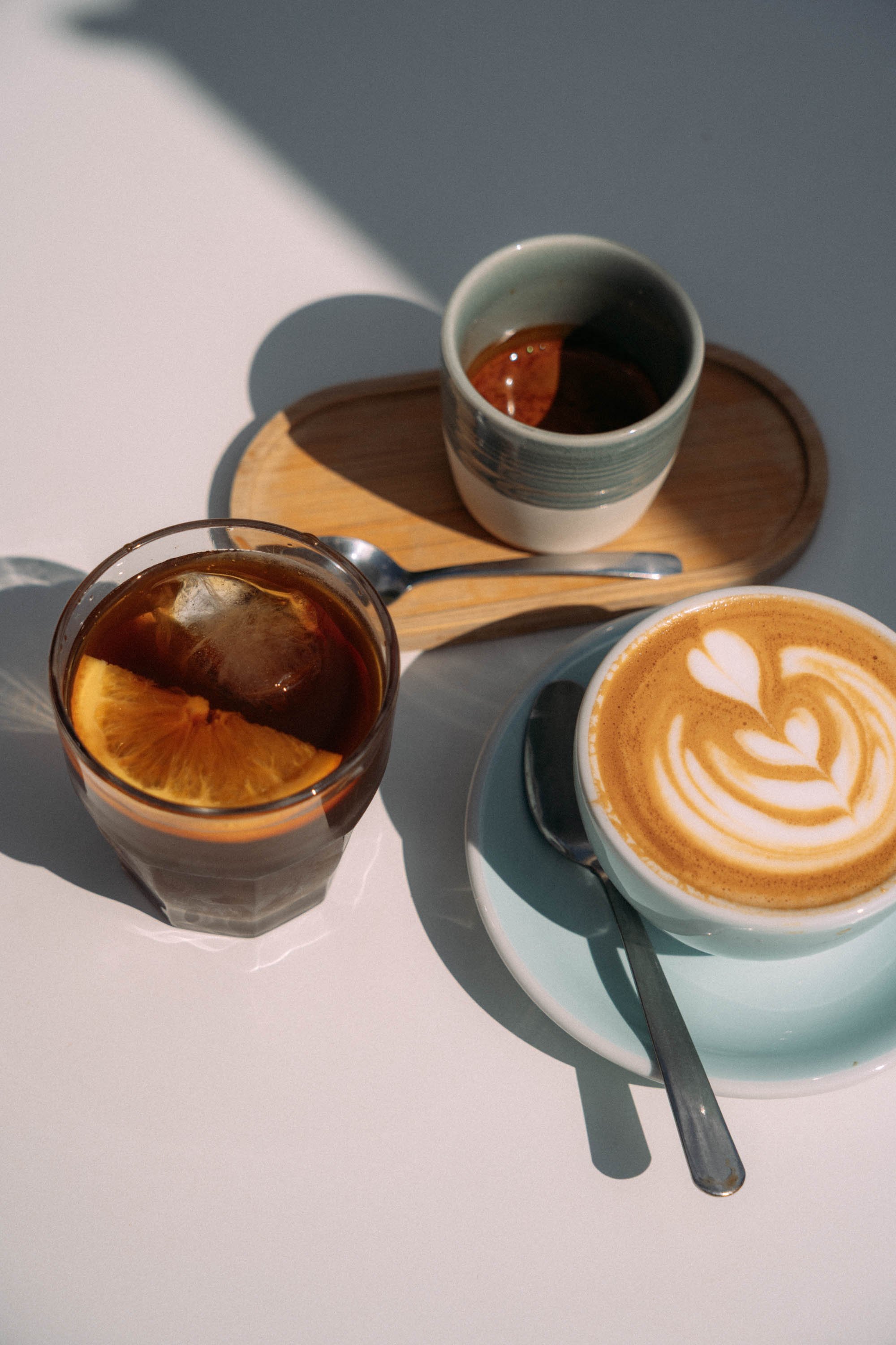 A top view of a glass of iced coffee with a slice of orange, a latte with latte art in a cup and saucer, and a ceramic cup of black coffee on a wooden tray, with a teaspoon.