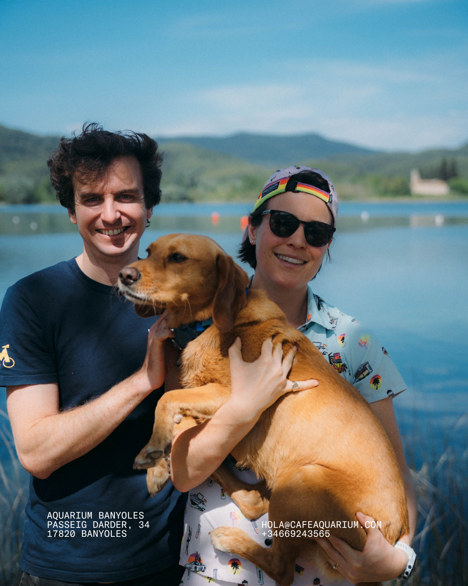 Two smiling people, a man with curly dark hair and a woman wearing sunglasses and a hat, are holding a large brown dog outdoors near a lake with mountains in the background.