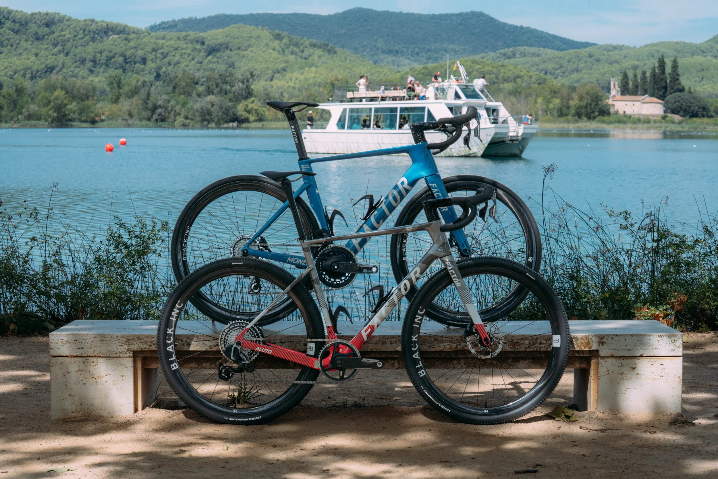 Two modern road bikes, one blue and one gray, are parked on a concrete bench next to a lake with a paddle wheel boat and green hills in the background.