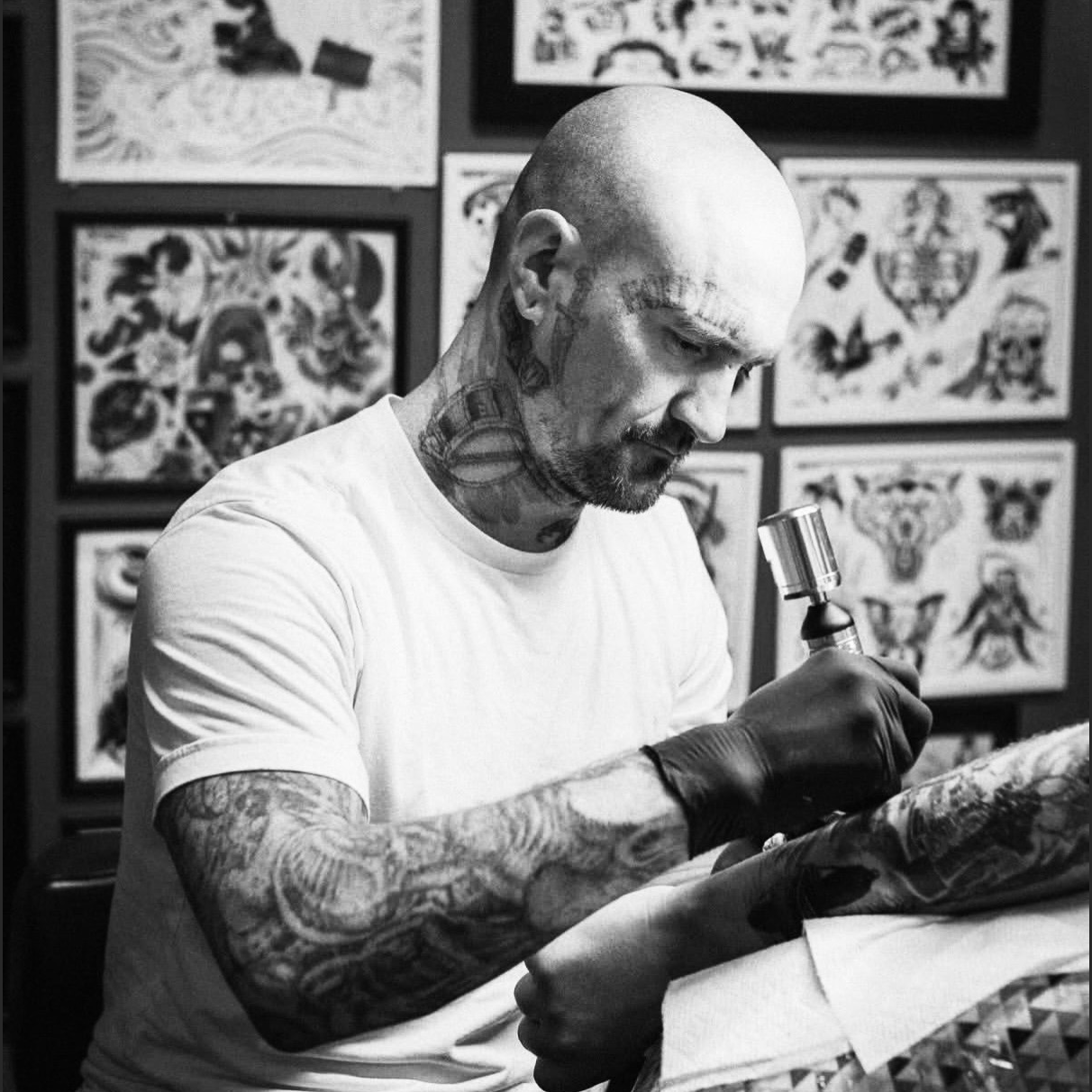 A man with tattoos on his head, neck, and arms is tattooing a client in a tattoo studio. The background features framed tattoo designs.