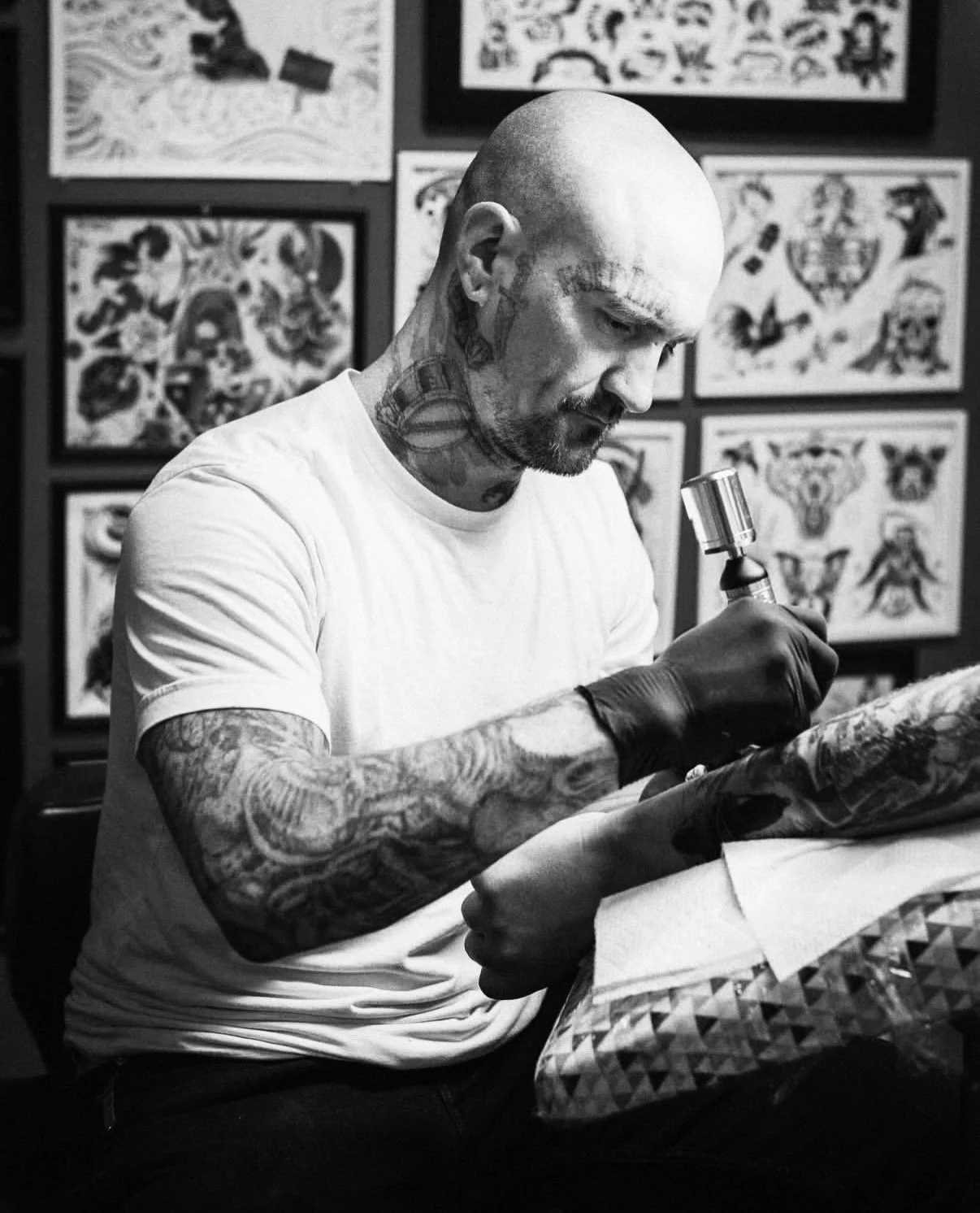 A tattooed man with a shaved head and beard, wearing a white t-shirt and gloves, performing a tattooing session with a tattoo machine in a tattoo studio. Tattoo flash art is displayed on the wall in the background.