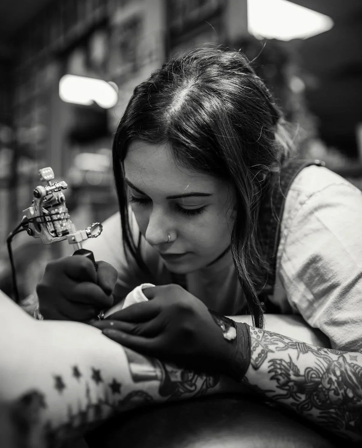 A woman with long dark hair tattooed arms, wearing gloves, a nose ring, and earrings, is tattooing another person in a tattoo studio. The environment is softly lit, with a blurred background.