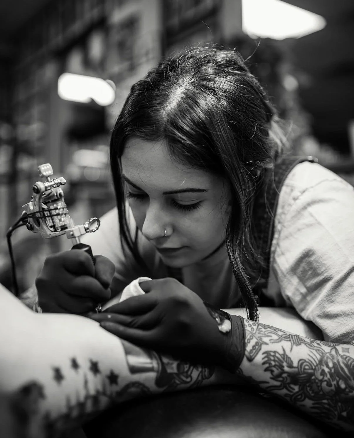A woman with dark hair, multiple tattoos, and a nose piercing is focused on tattooing a person's arm using a tattoo machine, in a tattoo studio environment.