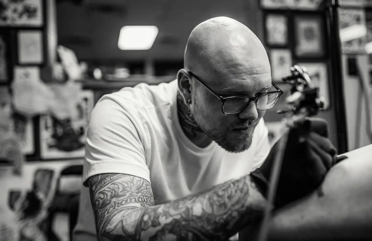 A tattooed man with a shaved head and glasses working on a tattoo machine in a tattoo studio.
