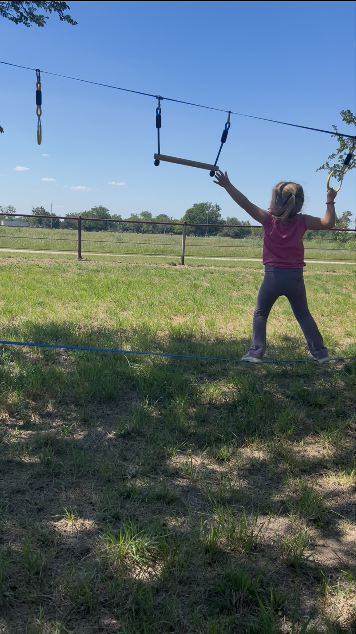 A young girl with blonde hair and a headband wearing a pink shirt and gray pants stands in a grassy field, reaching up toward a set of outdoor playground swings hanging from a wire, with a fence and trees in the background on a sunny day.