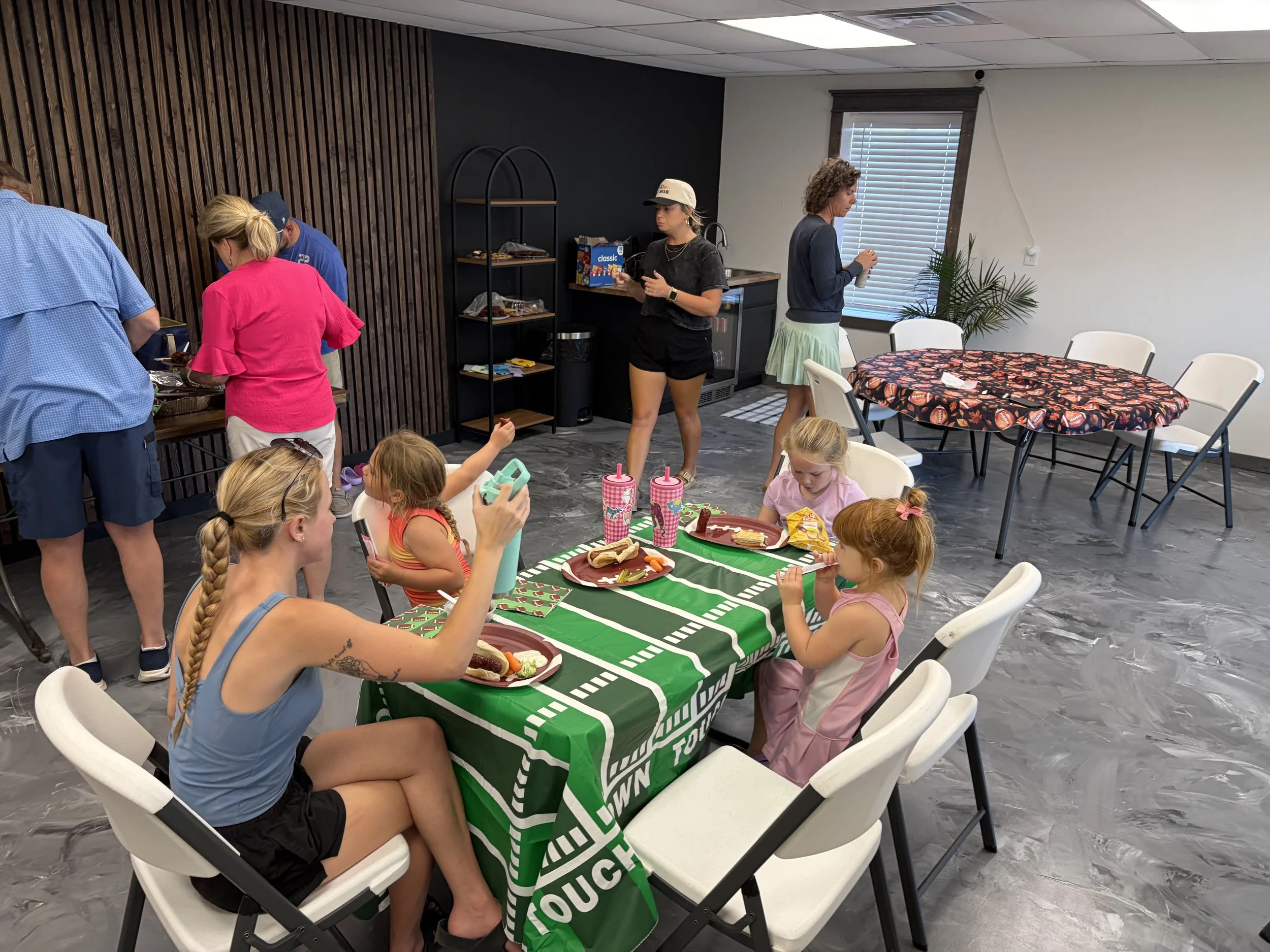 Children and adults gather around a table with food at an indoor party, some children sitting and some adults standing near the food table.