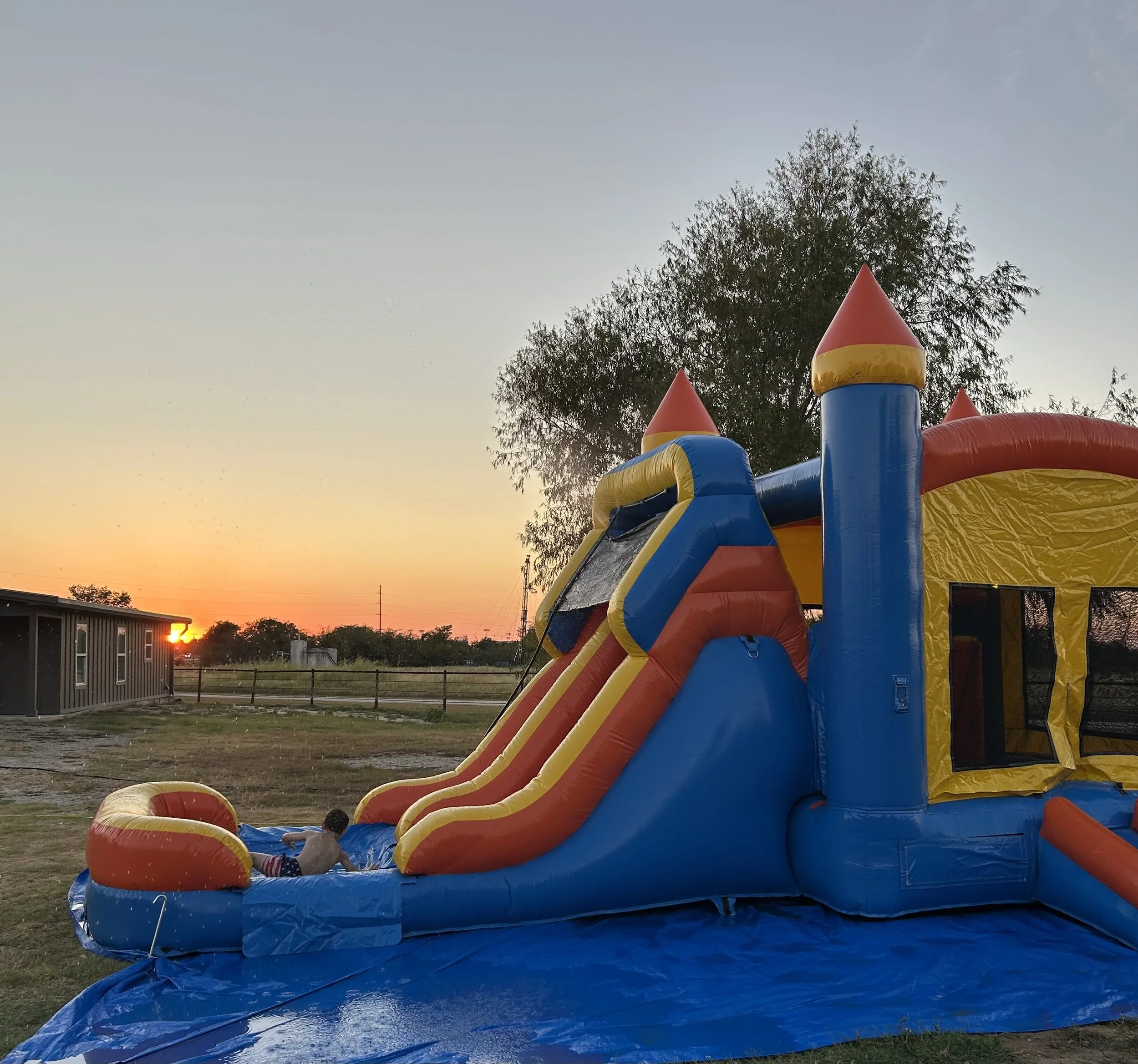 Colorful inflatable water slide on grass with trees in the background during sunset.