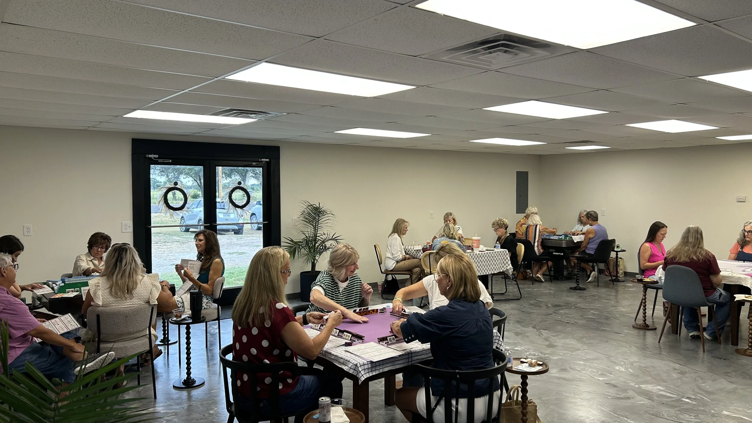 People gathered in a community room playing board games, socializing, and enjoying food, with large windows and plants inside.