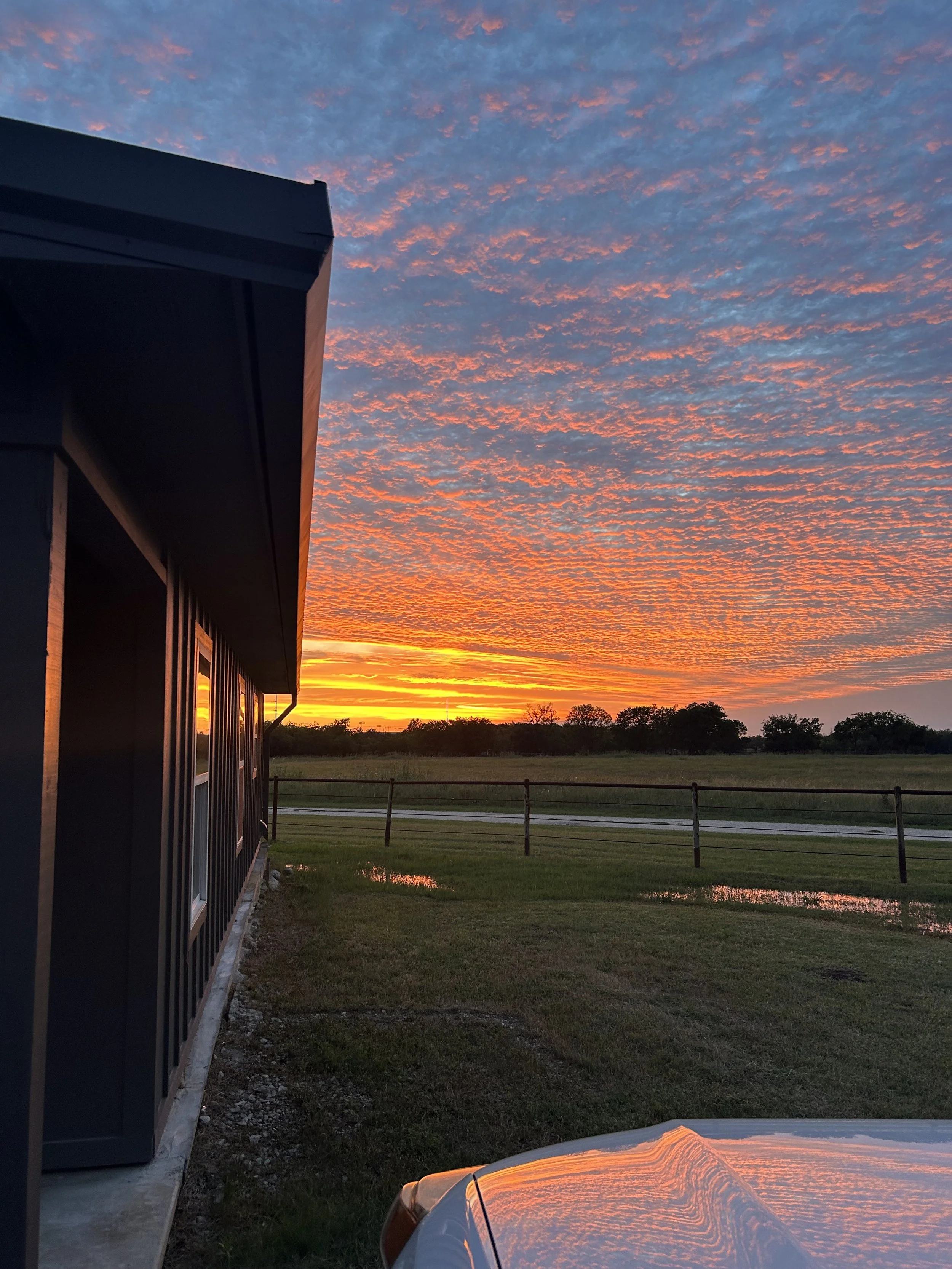 Sunset sky with vibrant orange and pink clouds over a rural landscape, with part of a building and a car's hood reflecting the sunset.