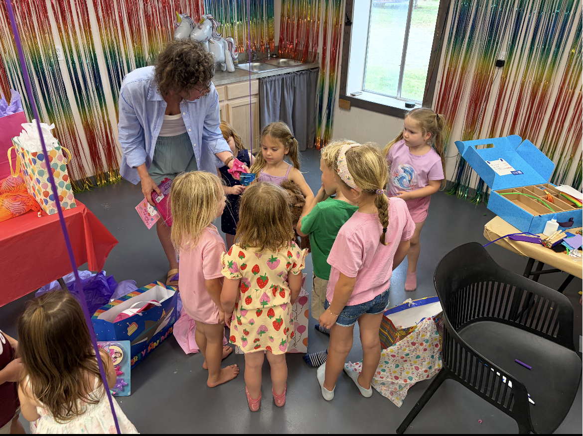 A group of young children gathered around a woman at a birthday party. The children are wearing colorful clothes with some in pink, as they look at presents or an activity. The room has rainbow streamers, a table with gifts, and a window showing a grassy outdoor area.