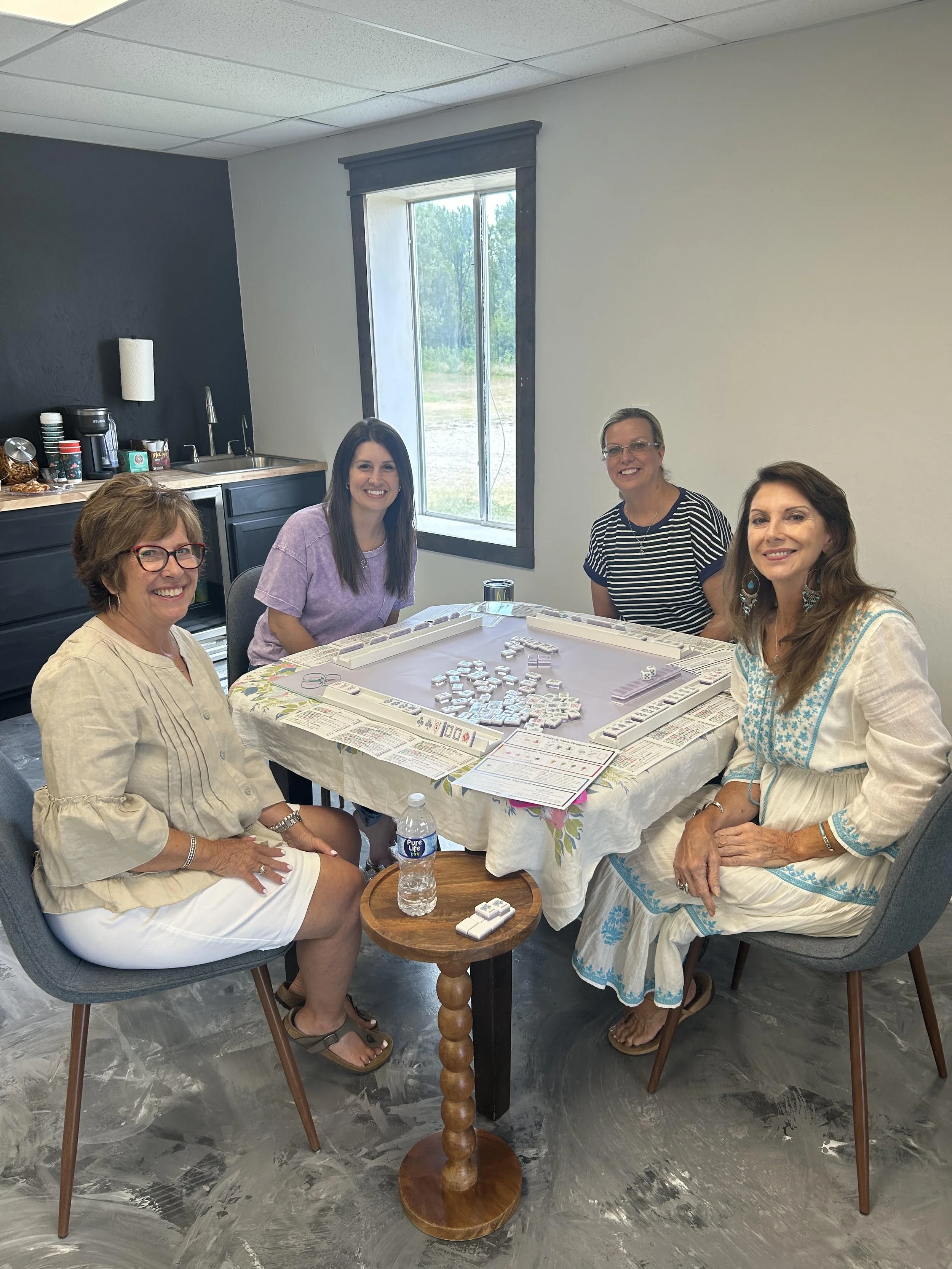Four women sitting around a table playing Mahjong in a room with a black and white wall, a window, and a kitchen counter with coffee and snacks.