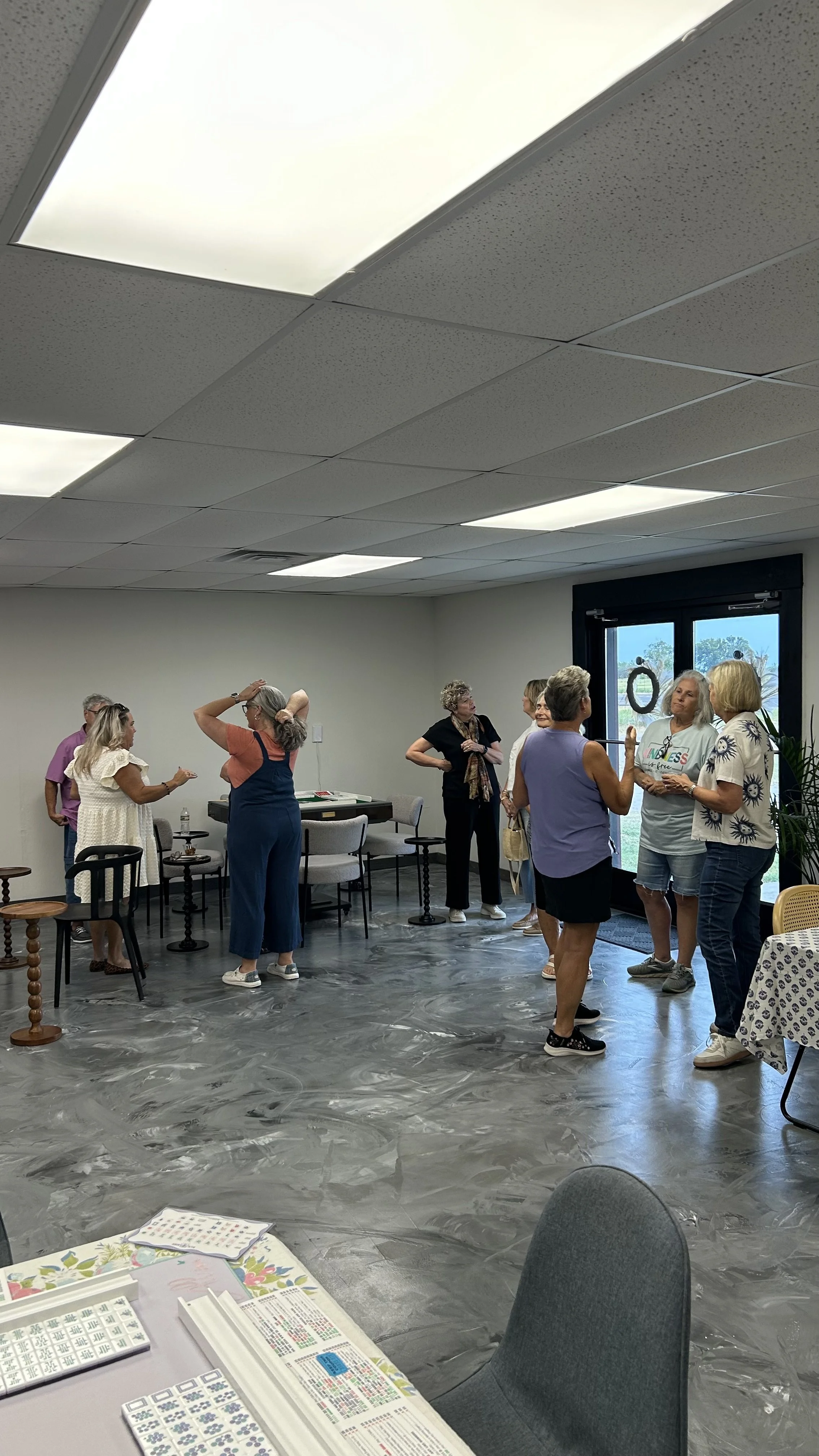 Group of people socializing and talking indoors in a room with a terrazzo floor, acoustic ceiling tiles, and large windows.