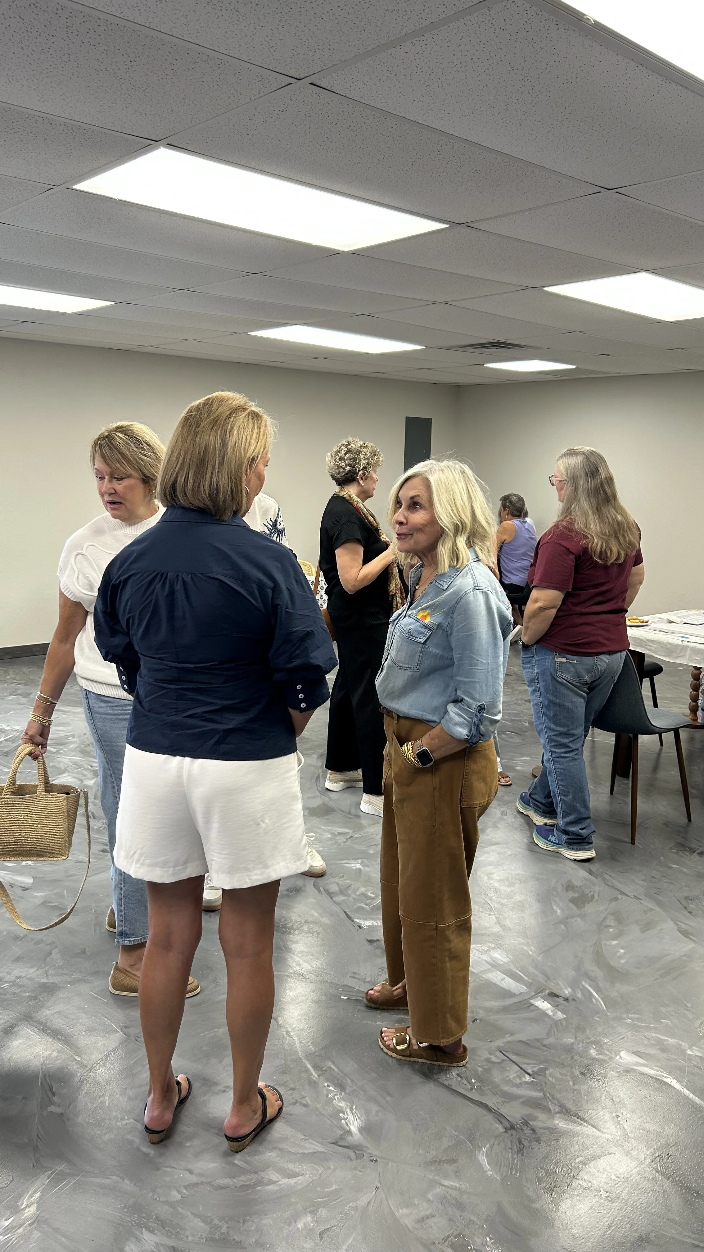 Group of women engaging in conversation in an indoor setting with a gray floor, plain gray walls, and fluorescent ceiling lights.
