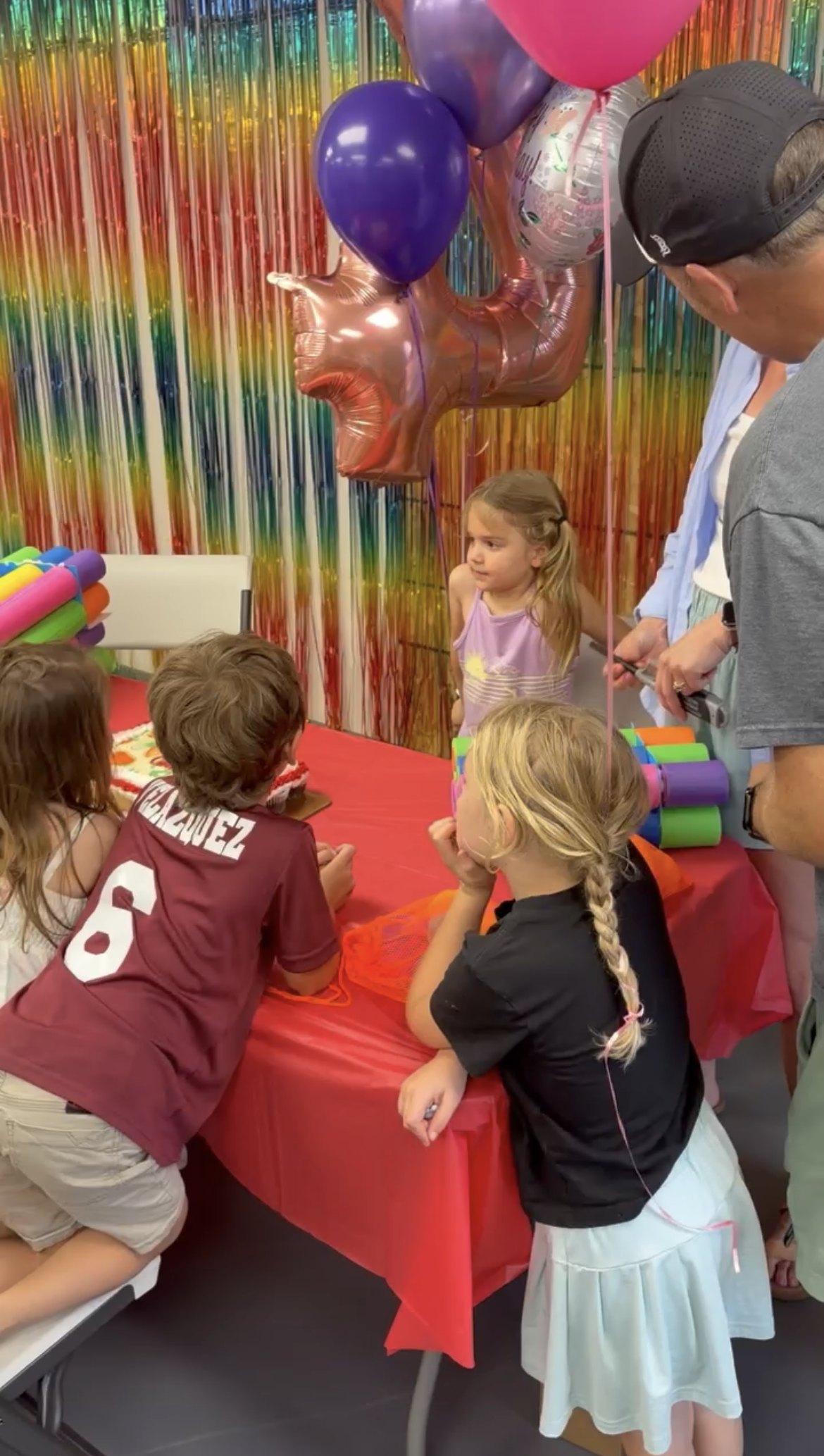 Children gathered around a table celebrating a birthday, with balloons, colorful decorations, and a rainbow backdrop.