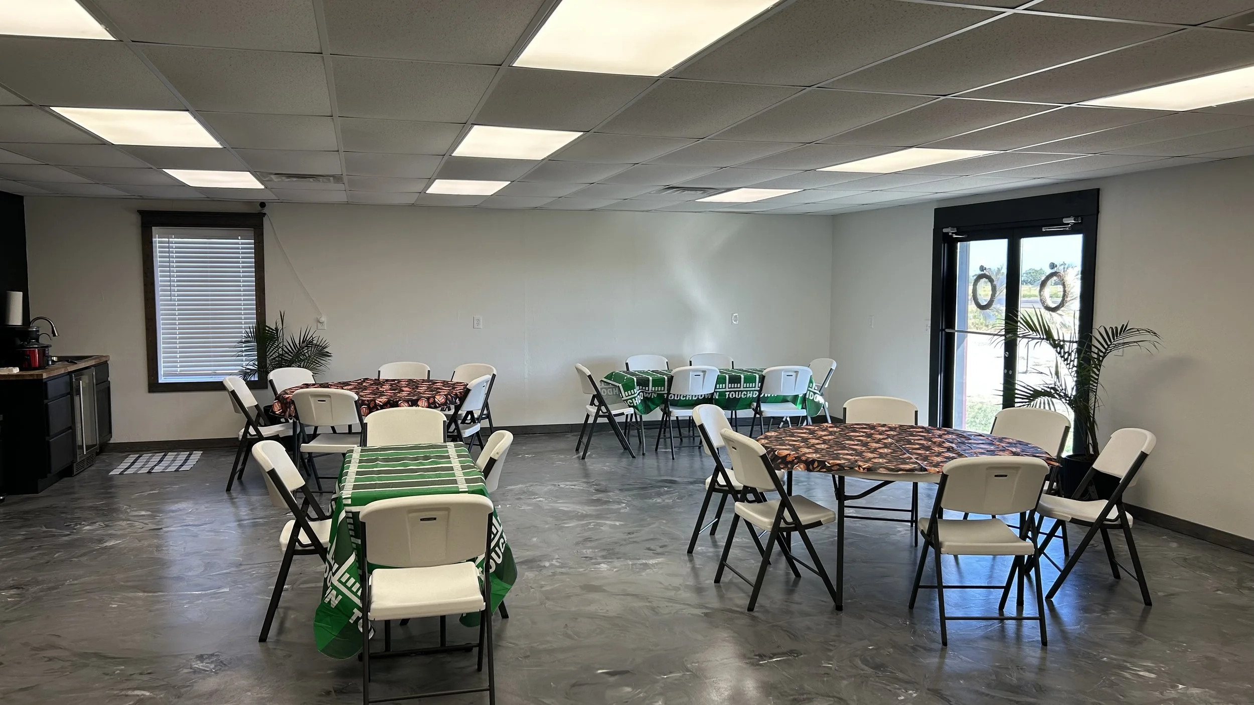 A room set up for a casual gathering with several round tables covered with green or floral tablecloths, surrounded by white chairs, with some tables having leftover plates, and large windows allowing natural light. There are plants and a small kitchenette area in the background.